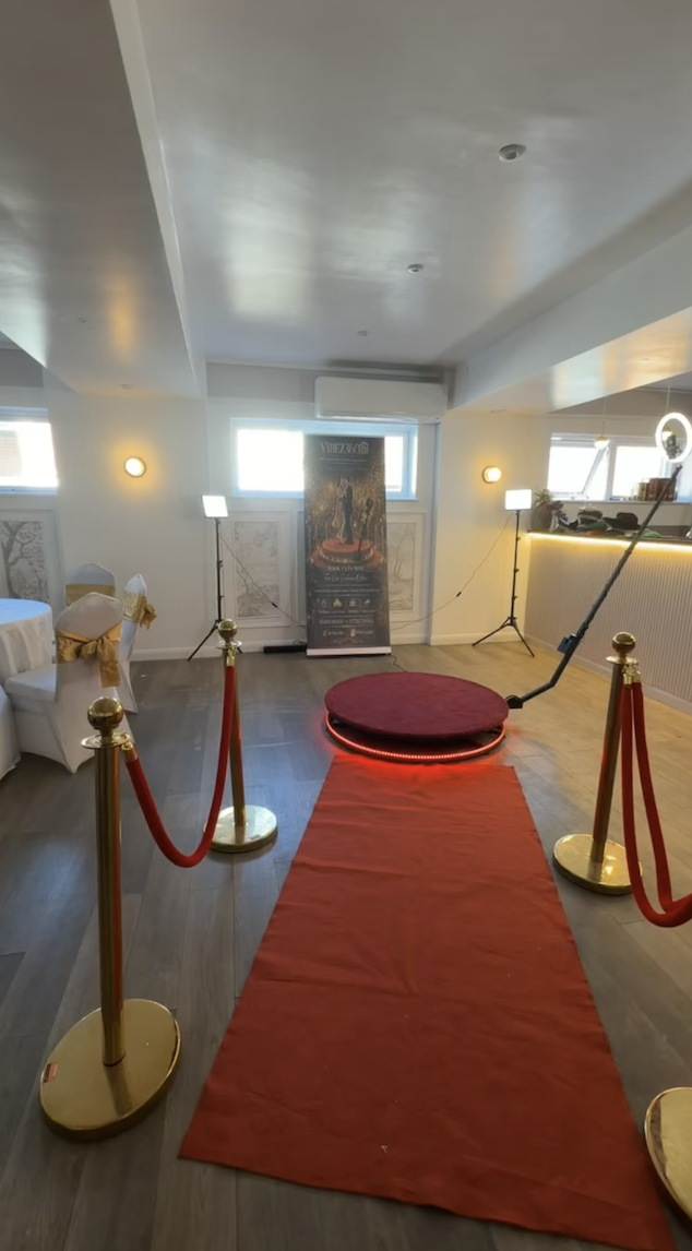 A vibrant party space features a red circular stage illuminated by multi-colored lights, gold balloons, and a DJ booth, creating a festive atmosphere.