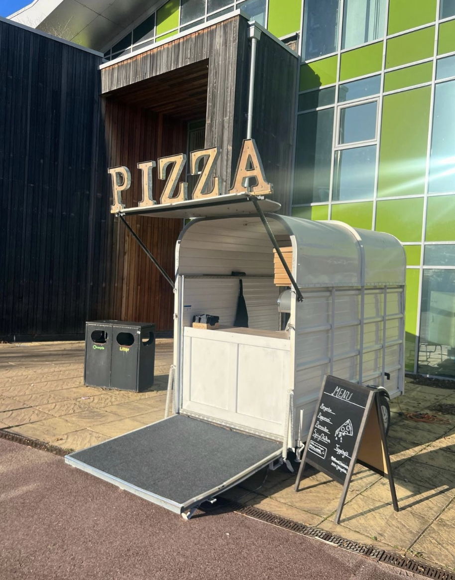 White pizza catering trailer with bold wooden sign, chalkboard menu and ramp, set before green-glass walls in bright daylight.