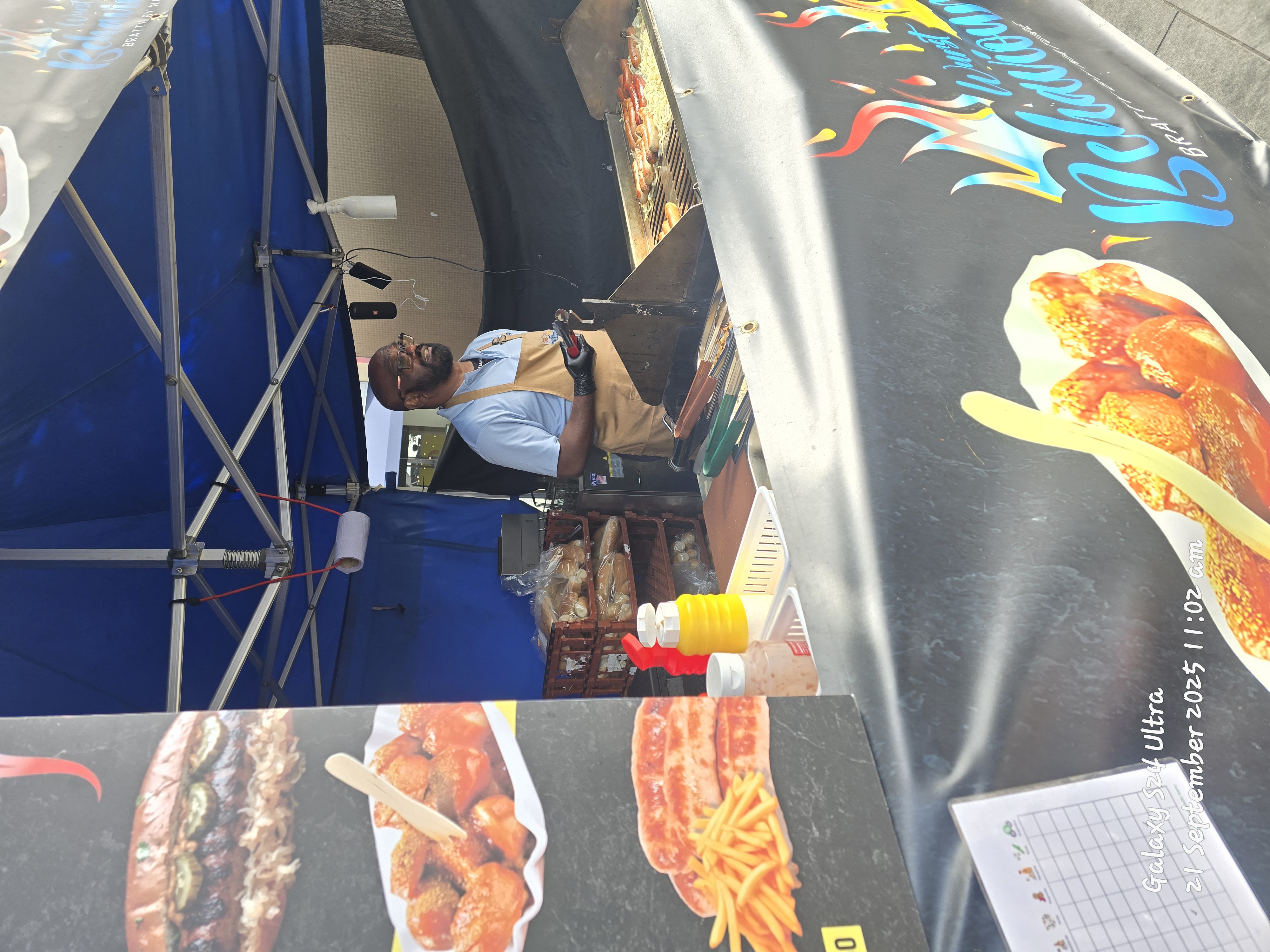 A lively food stall with a blue canopy features a smiling vendor grilling sausages, vibrant condiments, and a menu showcasing enticing dishes.