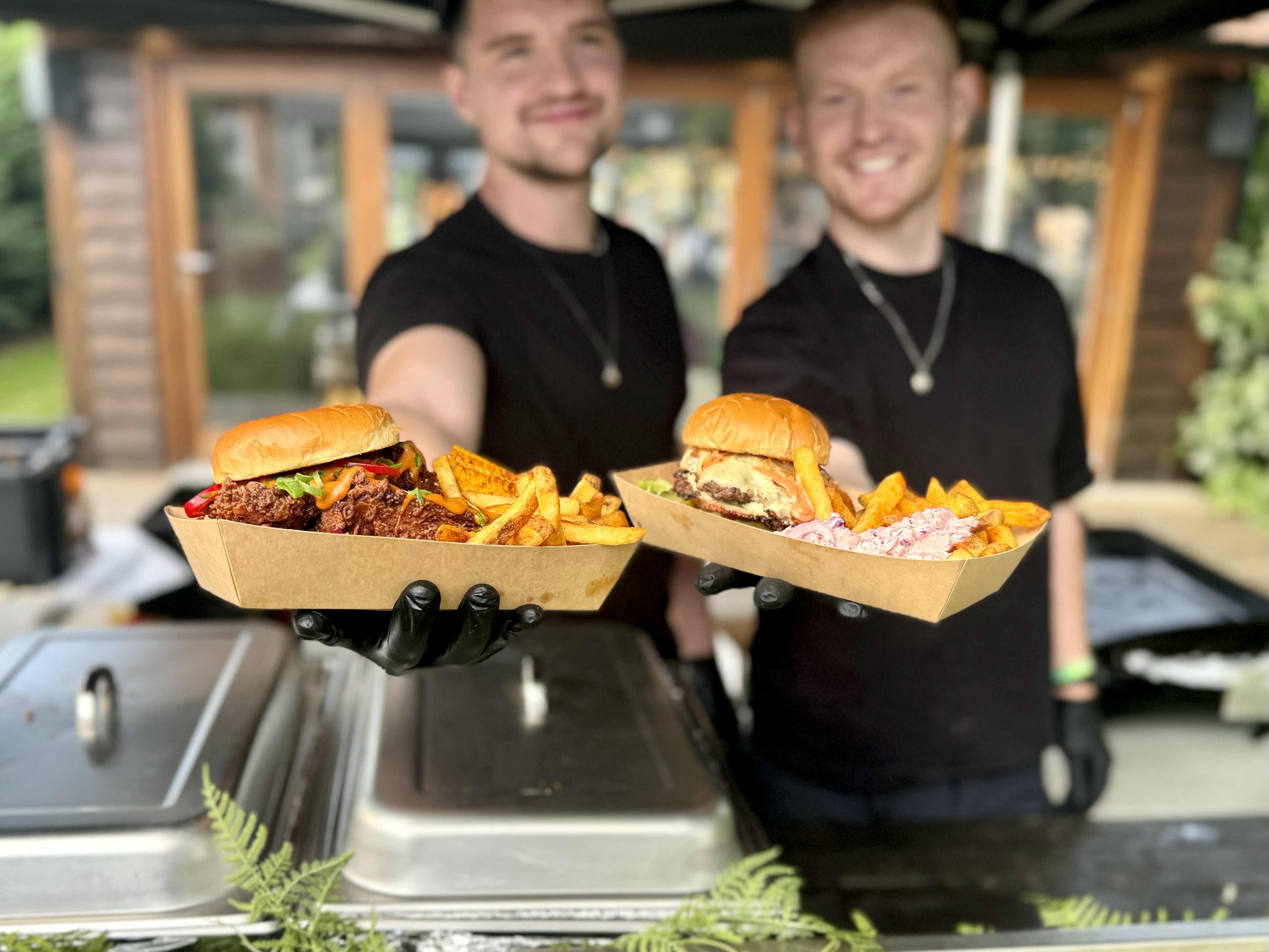 Two smiling chefs present colorful paper trays filled with vibrant burgers and golden fries, under a shaded outdoor catering setup.