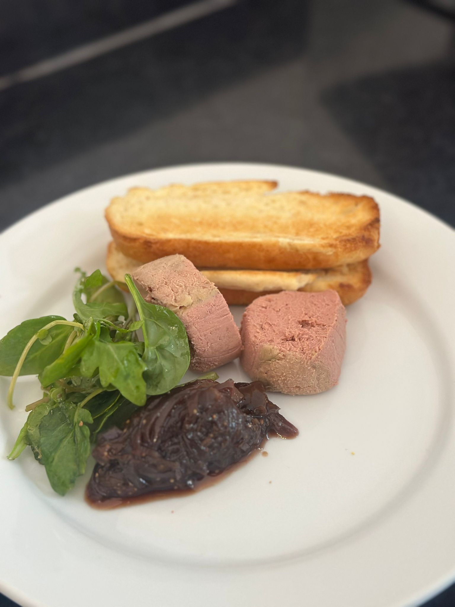 A pristine white plate holds three smooth pâté slices in pink tones, accompanied by crisp toasted bread and fresh greens, exuding elegance.