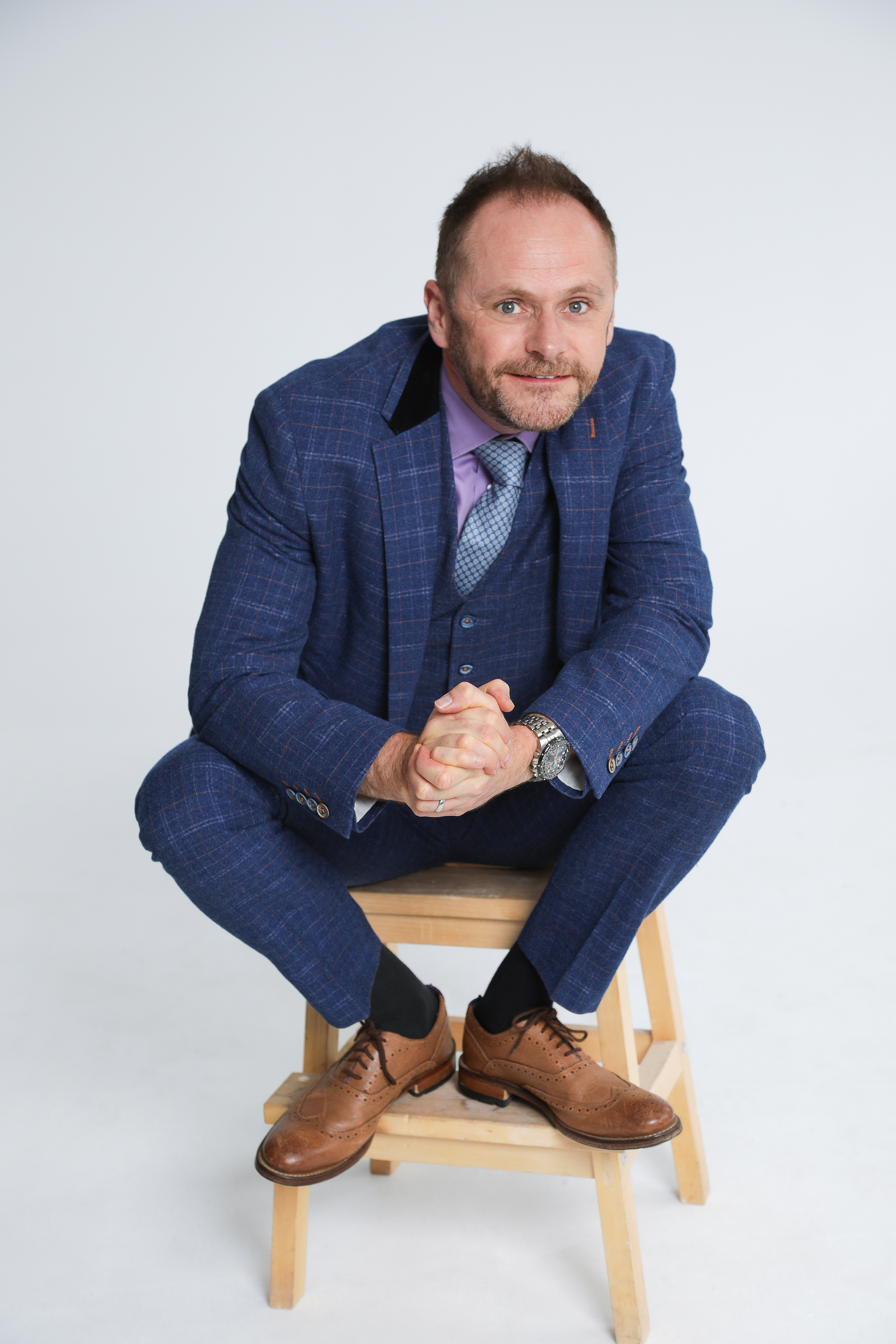 A man in a tailored blue suit and light shirt poses thoughtfully against a plain backdrop, conveying a professional, introspective vibe.
