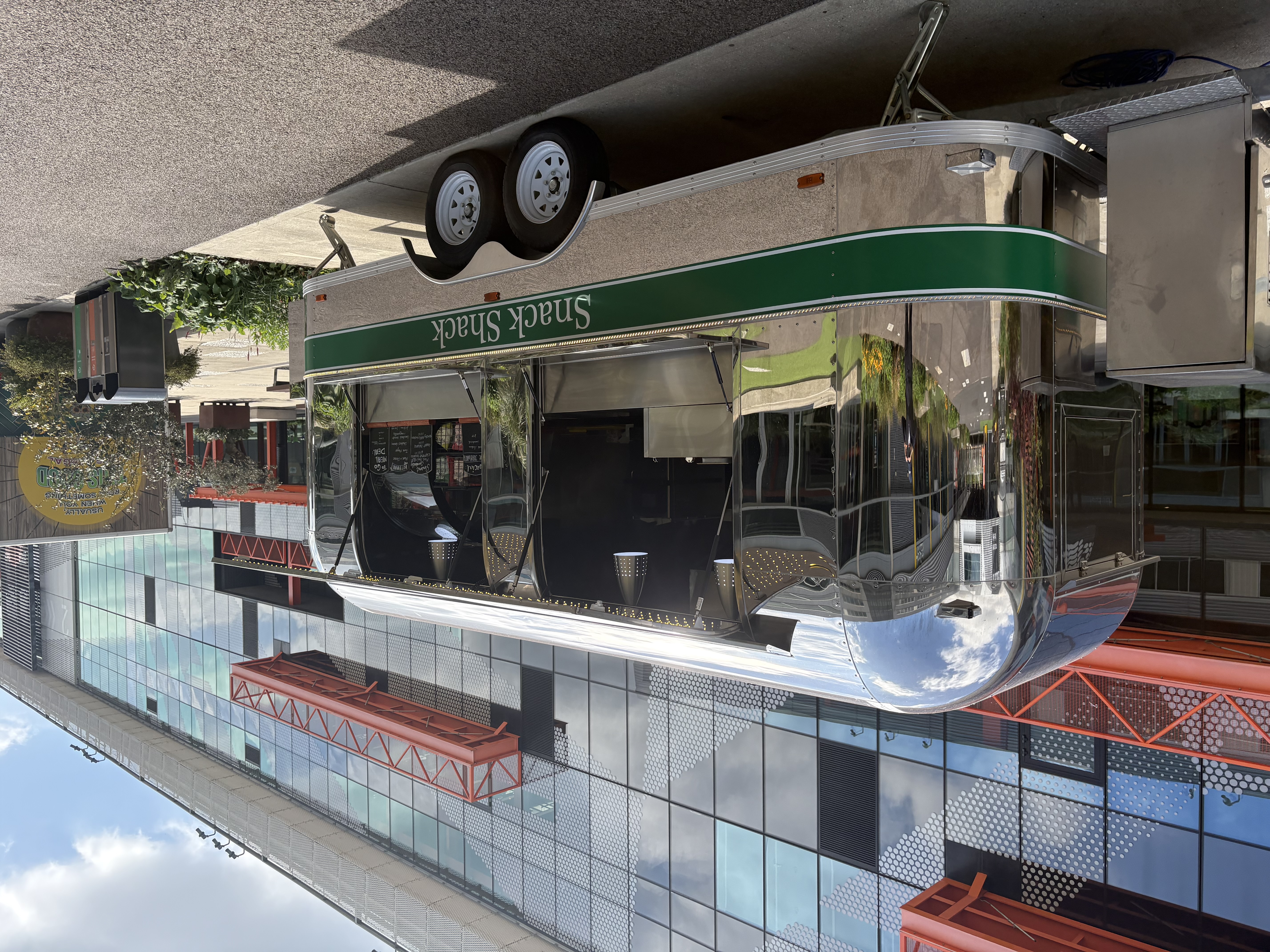 Polished chrome Snack Shack catering trailer with green trim and open service windows, parked by a glass venue under bright daylight.