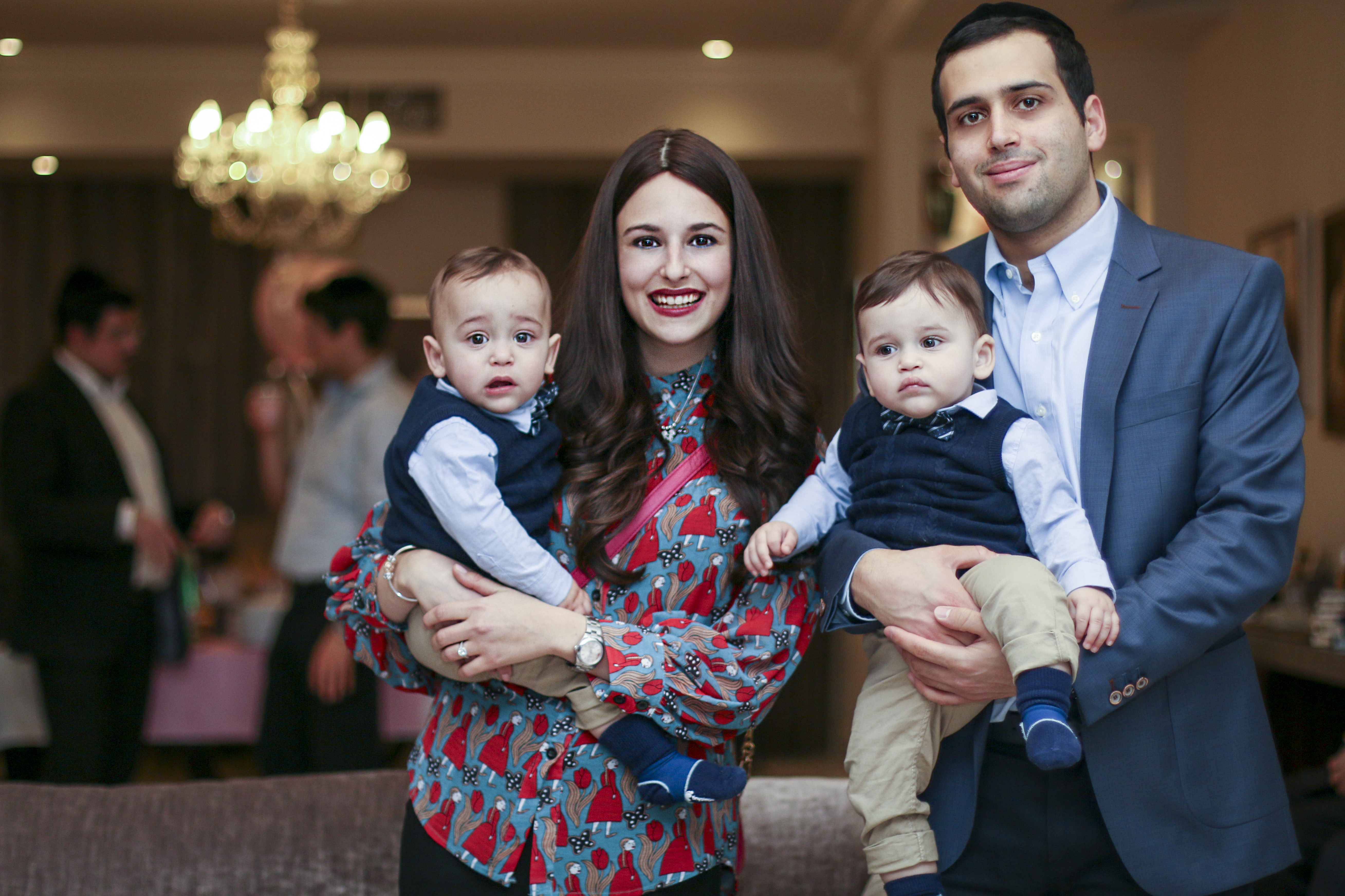 A smiling woman holds two toddlers, each in cozy vests, while a stylish man stands beside her. Soft chandelier light creates a warm ambiance.