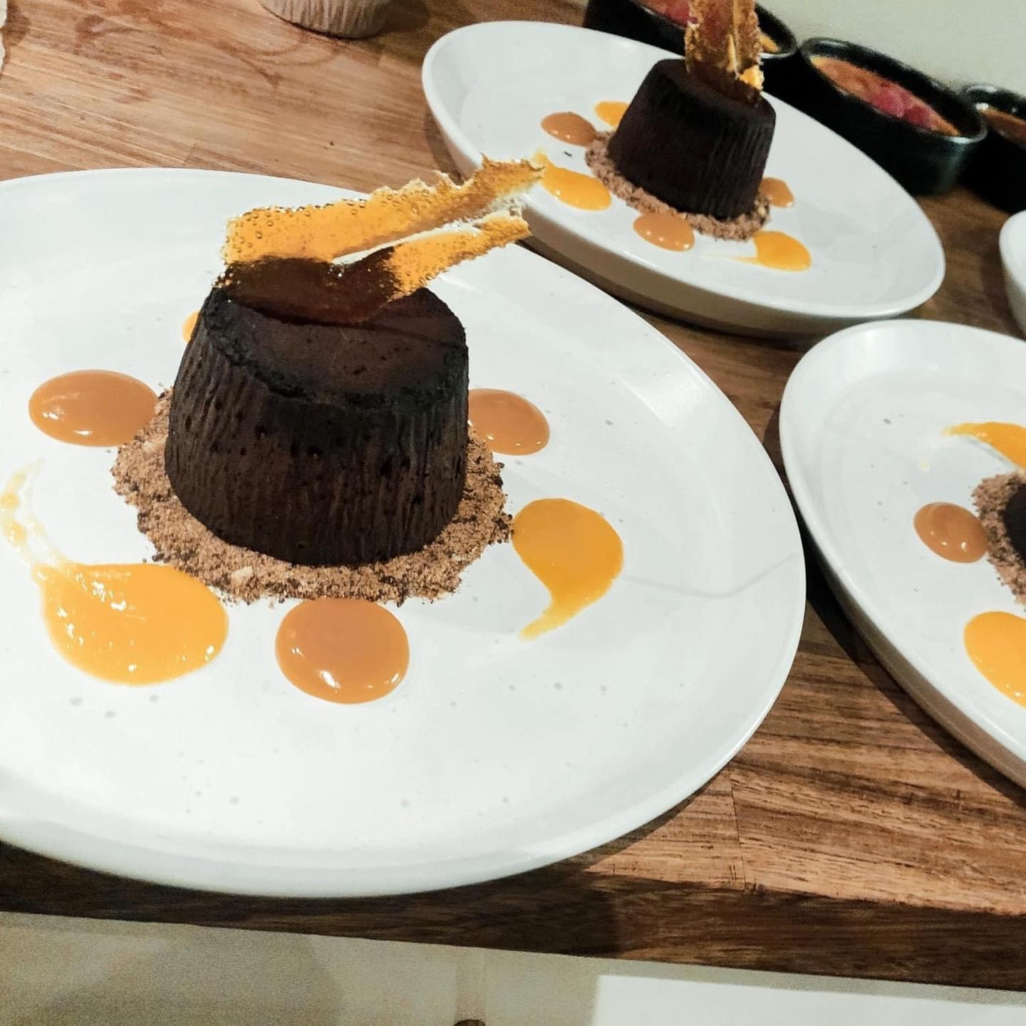Decadent chocolate desserts are elegantly arranged on white plates, surrounded by vibrant orange sauces and crumbly textures, enhancing the catering’s quality.