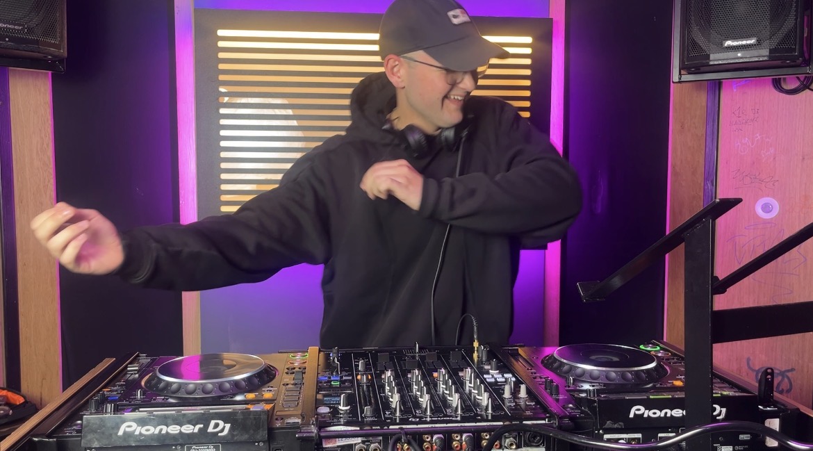 DJ at a Pioneer setup spins in purple-lit booth, with twin decks and mixer centered, clean club catering energy, polished pro sound.