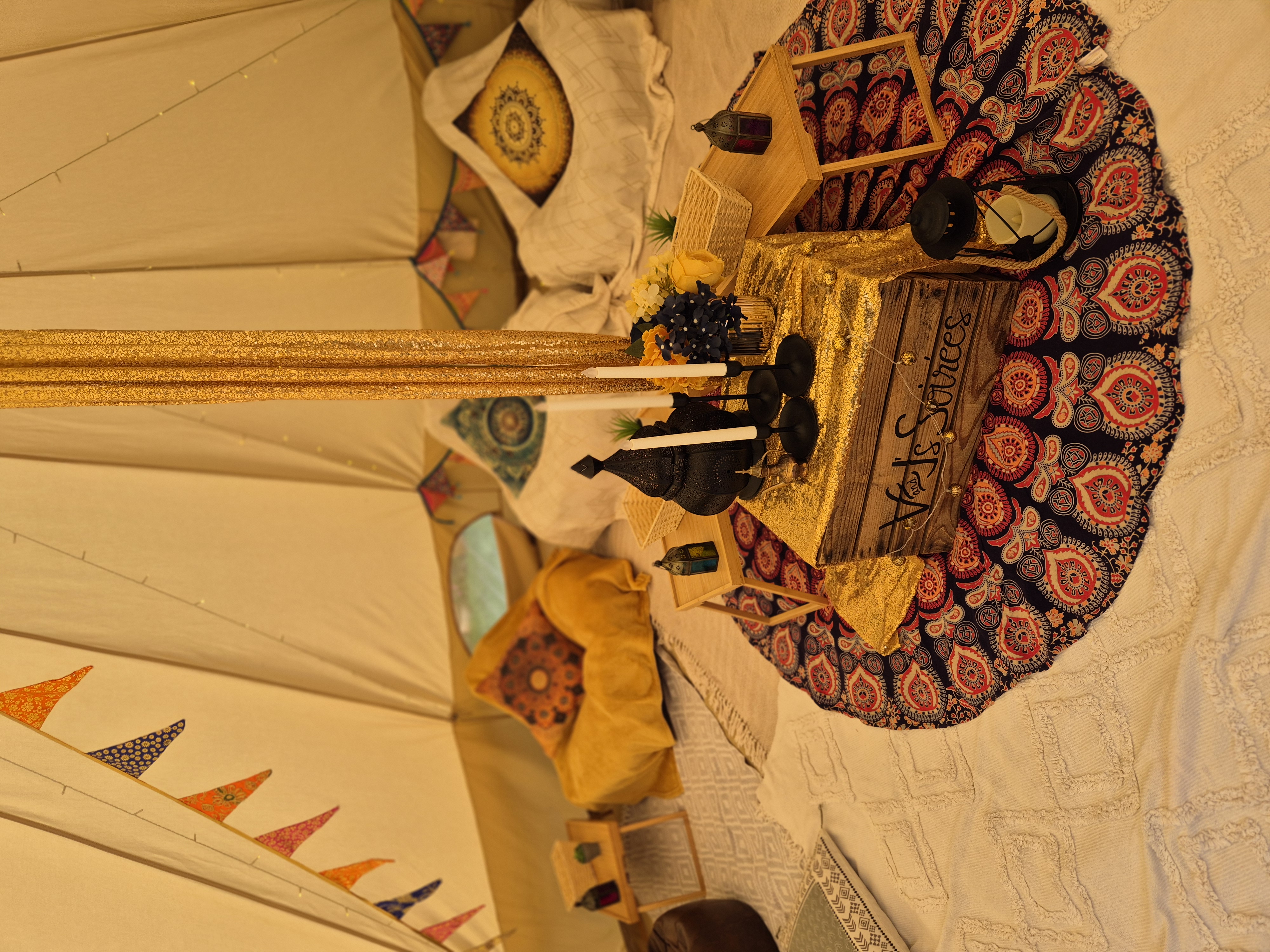 Warm-lit boho tent lounge with patterned cushions, gold drapes, lanterns, and a rustic wood centerpiece for stylish catering vibes.