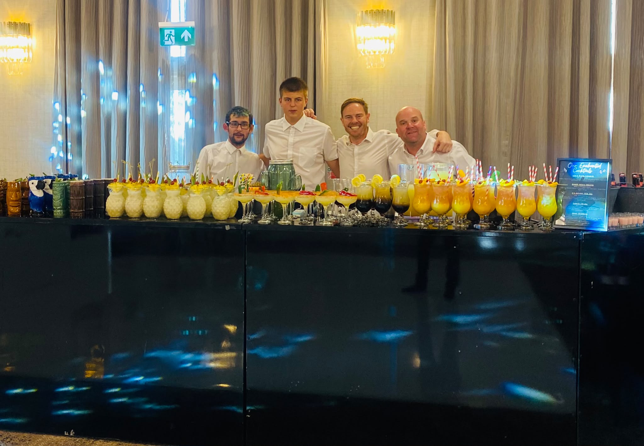 Smiling bartenders behind a sleek black catering bar, serving neatly lined tropical cocktails and lemon drinks under warm chandelier light.