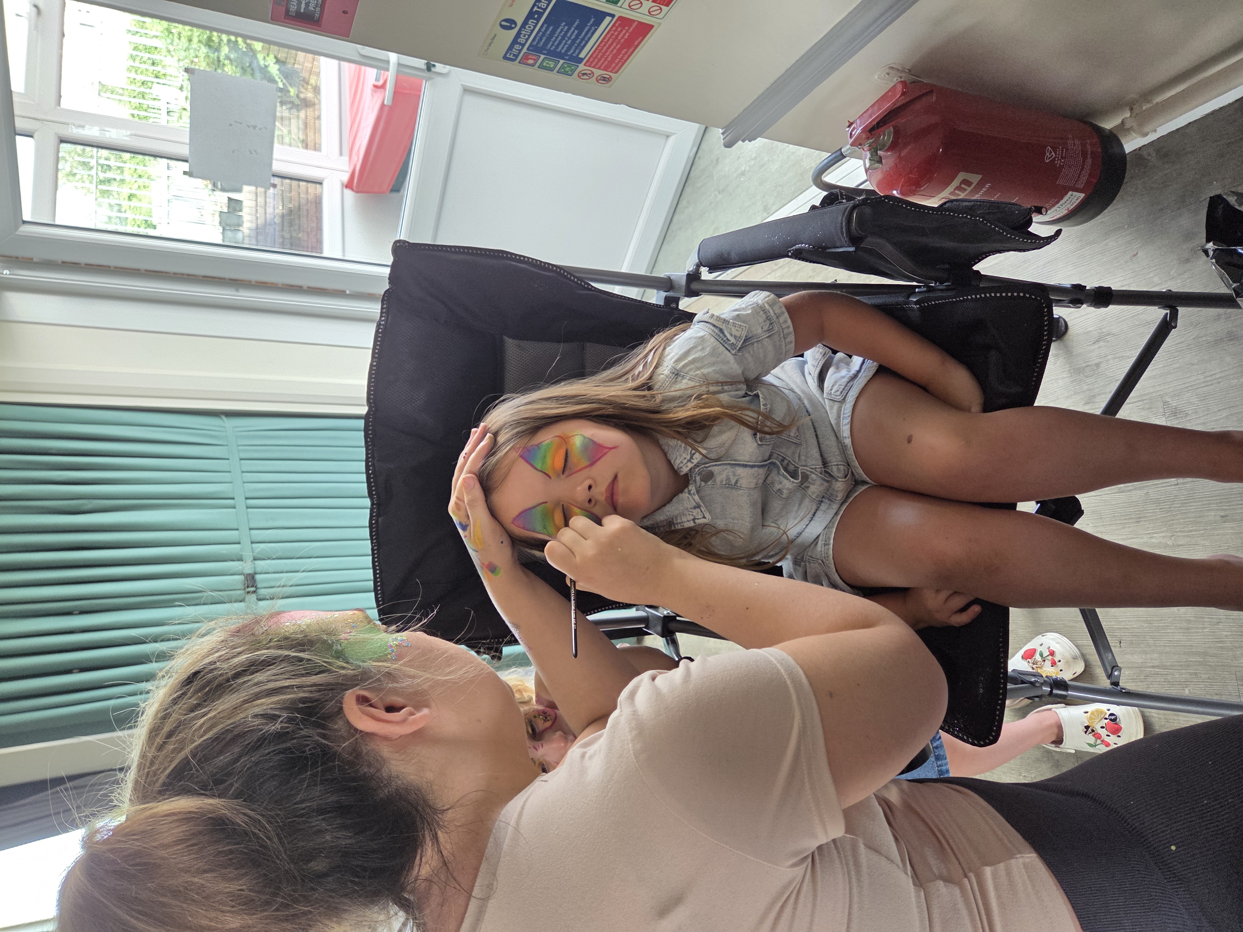 Face-painting service at a bright venue, with a child in a black chair and rainbow makeup, polished and cheerful.