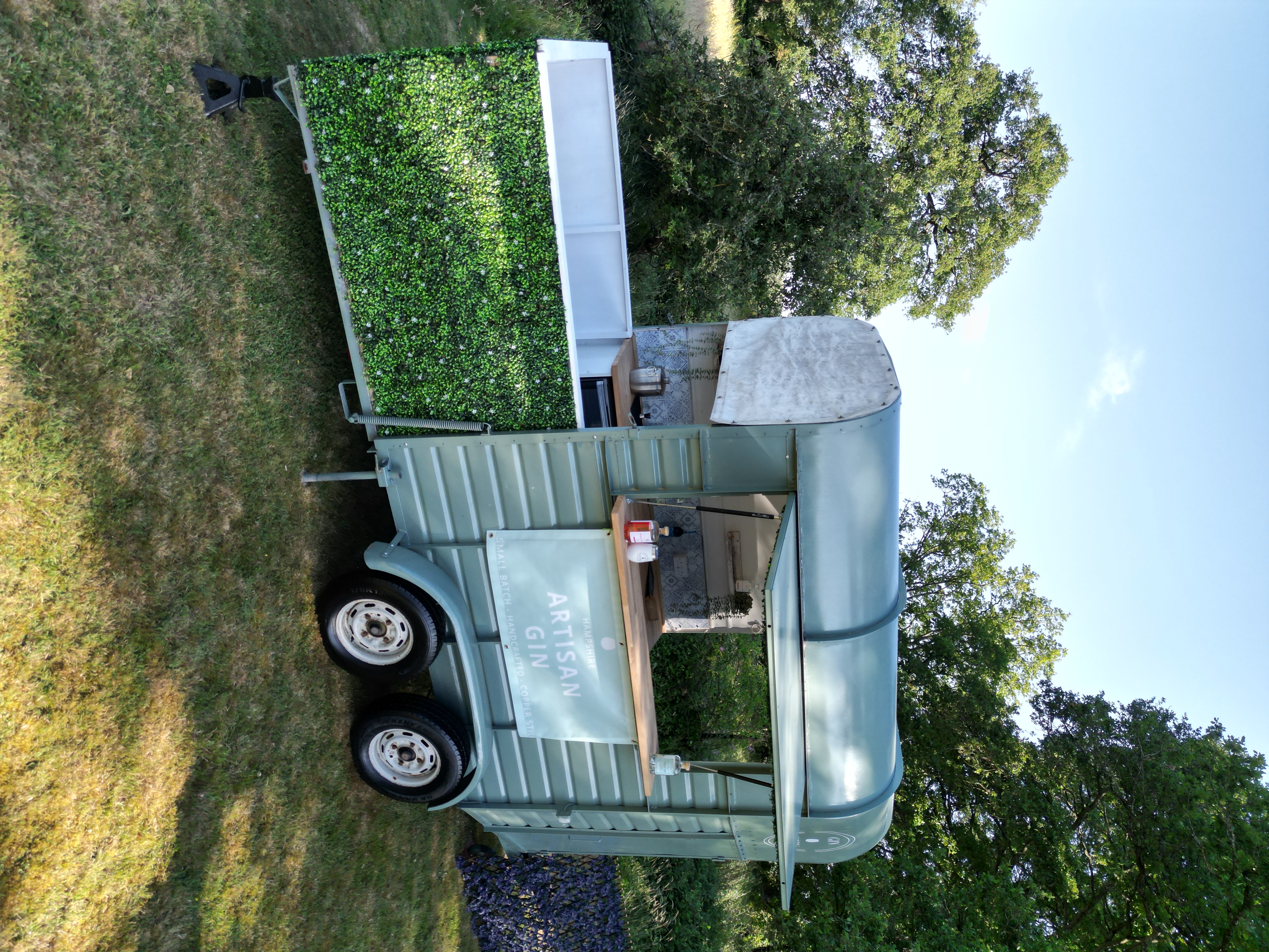 A vintage-style gin caravan features a rustic wooden bar, adorned with greenery, under bright daylight, showcasing a charming outdoor setup for catering.