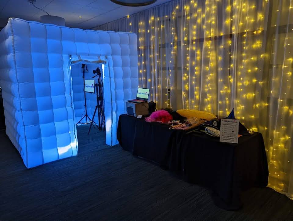 A cozy event setup features a bright blue photo booth, twinkling fairy lights, a black table with vibrant props, and warm ambient lighting.