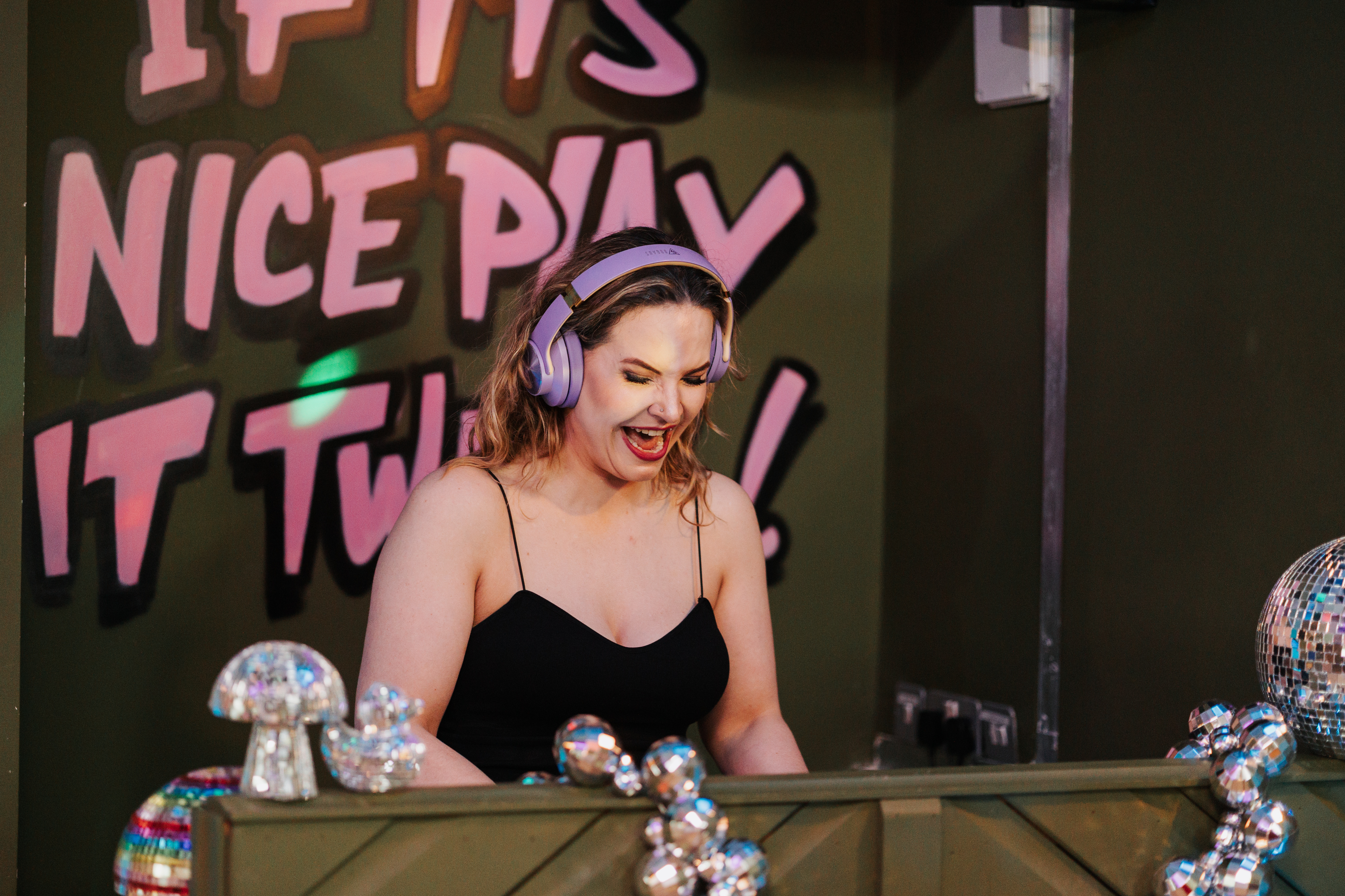 A DJ in a black tank top, sporting purple headphones, energetically mixes music against a vibrant green wall with playful graffiti.