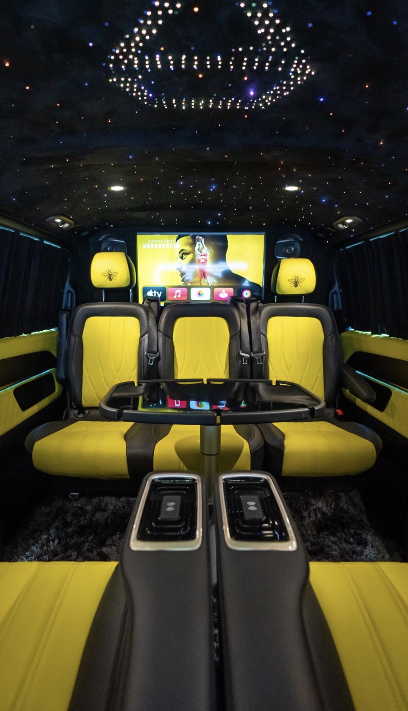 The luxurious interior features sleek black and vibrant yellow leather seats with a plush rug, accented by a starry ceiling and large screen.