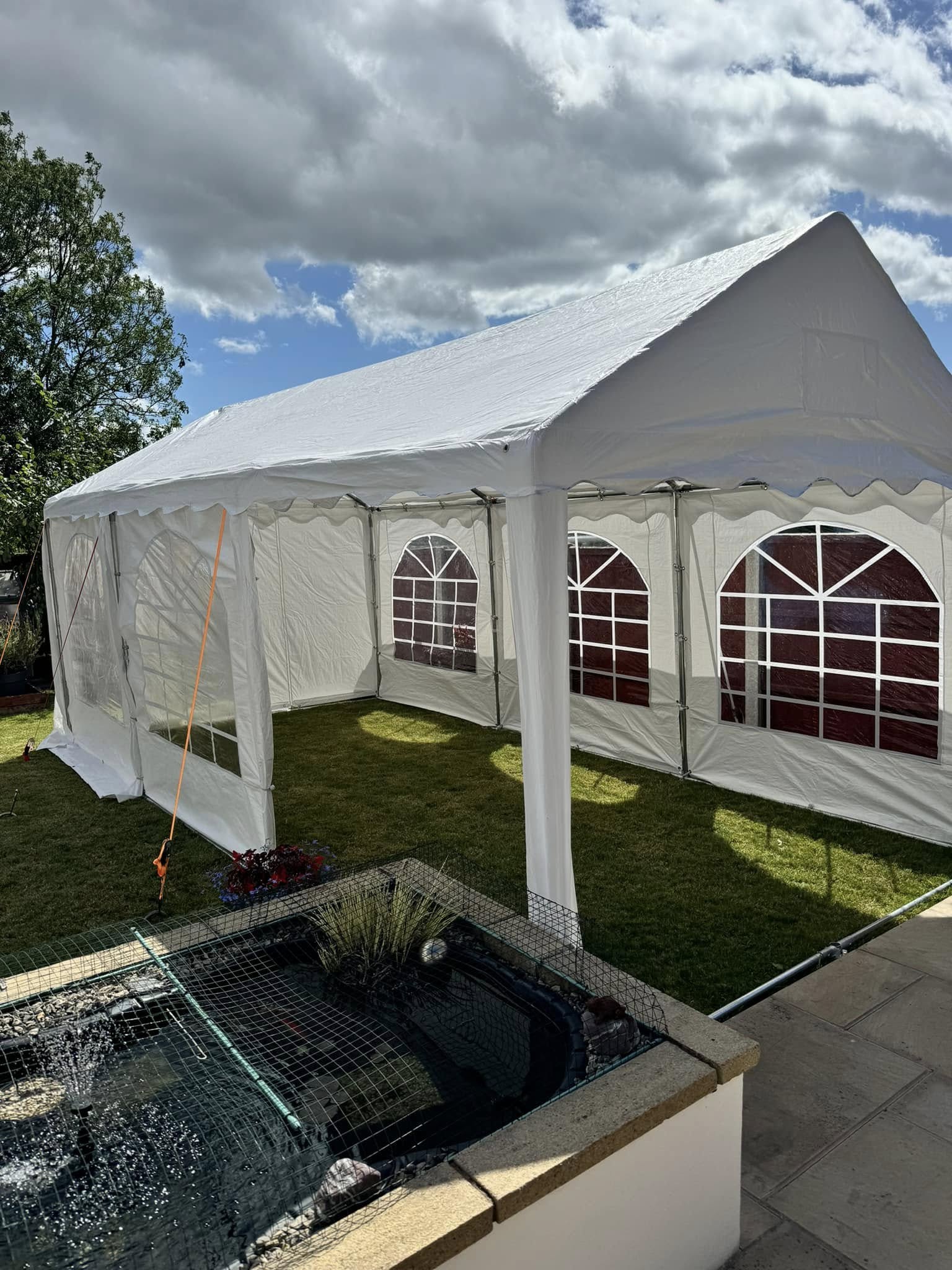 A spacious white tent with red-trimmed windows is set on lush green grass, next to a tranquil pond under bright daylight, perfect for events.
