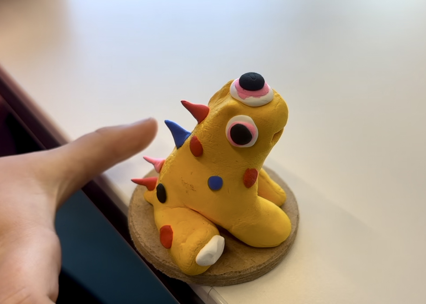 Handmade yellow clay monster with colorful spikes and googly eyes on a wooden base, lit by soft indoor daylight, artisanal display.