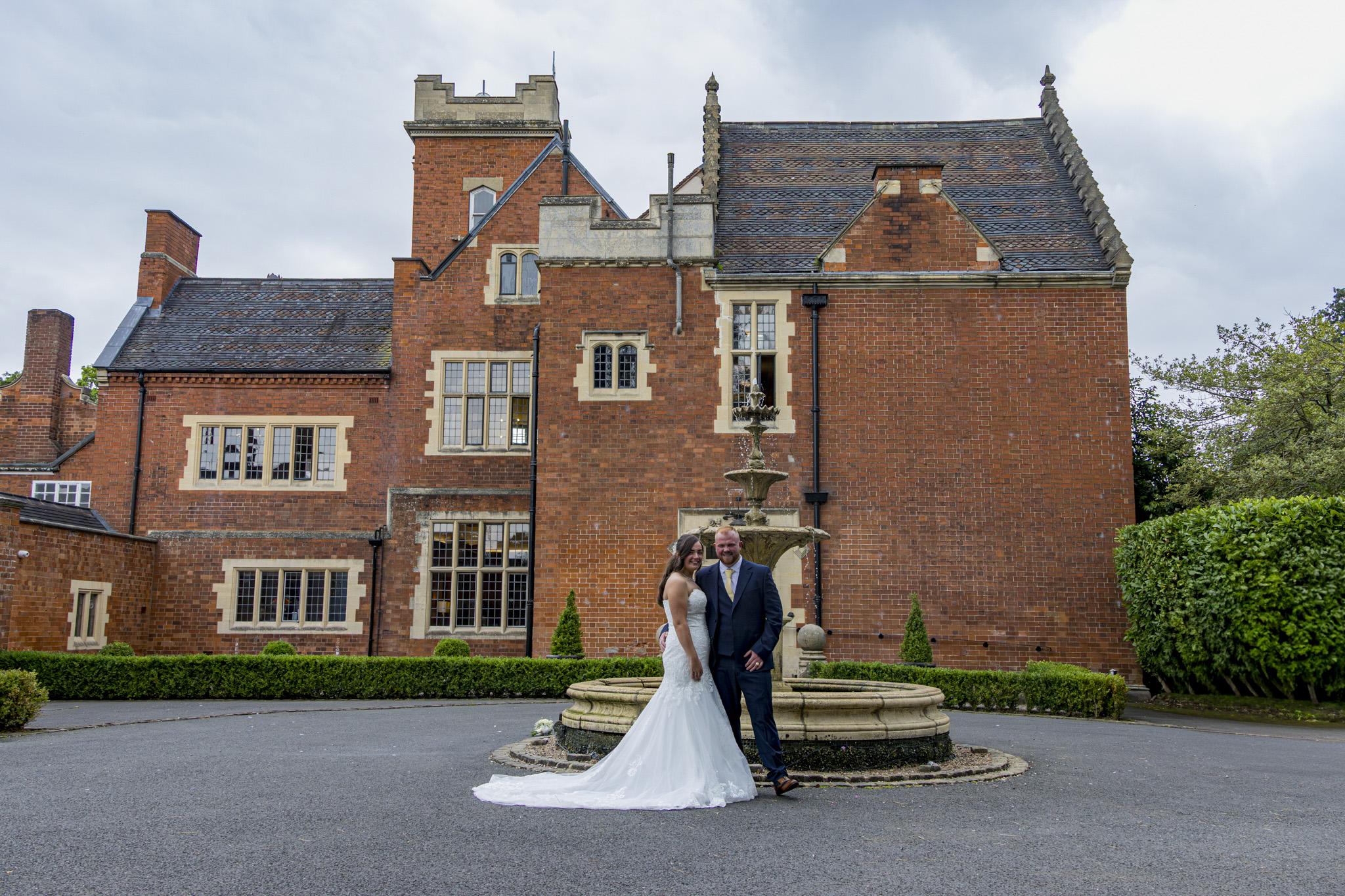 A couple stands by a charming stone fountain in front of a stunning red brick mansion, under soft, overcast skies, exuding romance.