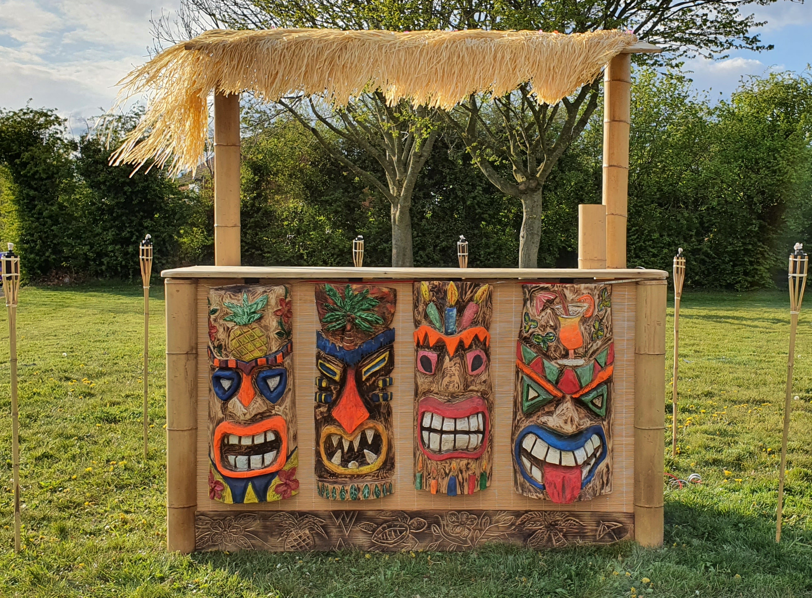 A vibrant tiki bar features colorful carved masks on a bamboo frame, topped with a thatched roof, set against a sunny garden backdrop.