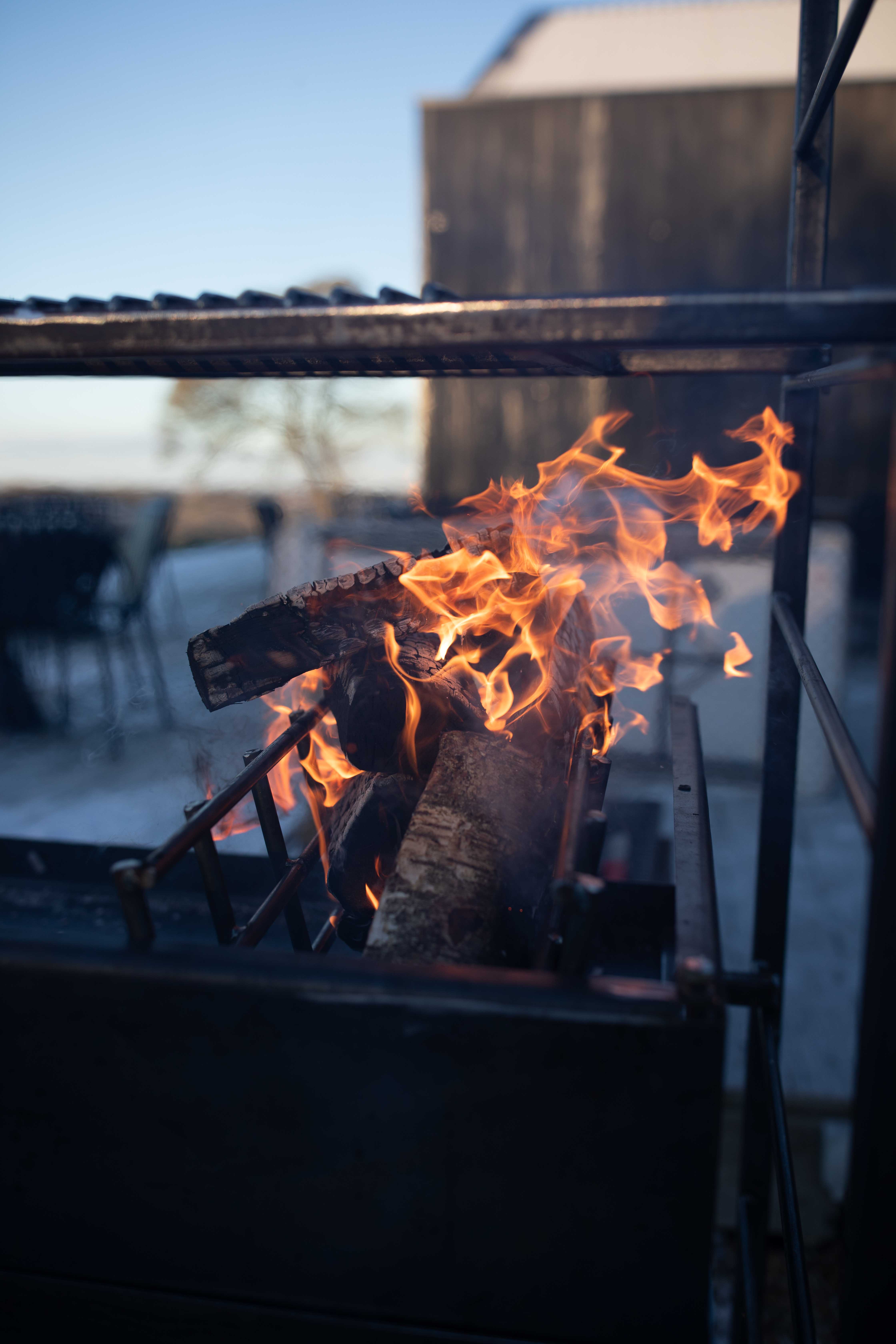 Open-air catering grill with stacked logs blazing in a black steel firebox, warm orange flames, cool blue sky, crisp evening ambiance.