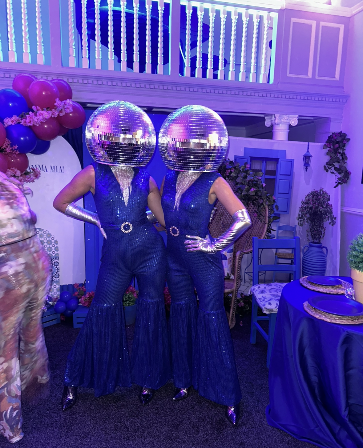 Two performers in dazzling blue sequined jumpsuits and silver gloves sport mirrored disco ball helmets, against a lively event backdrop.