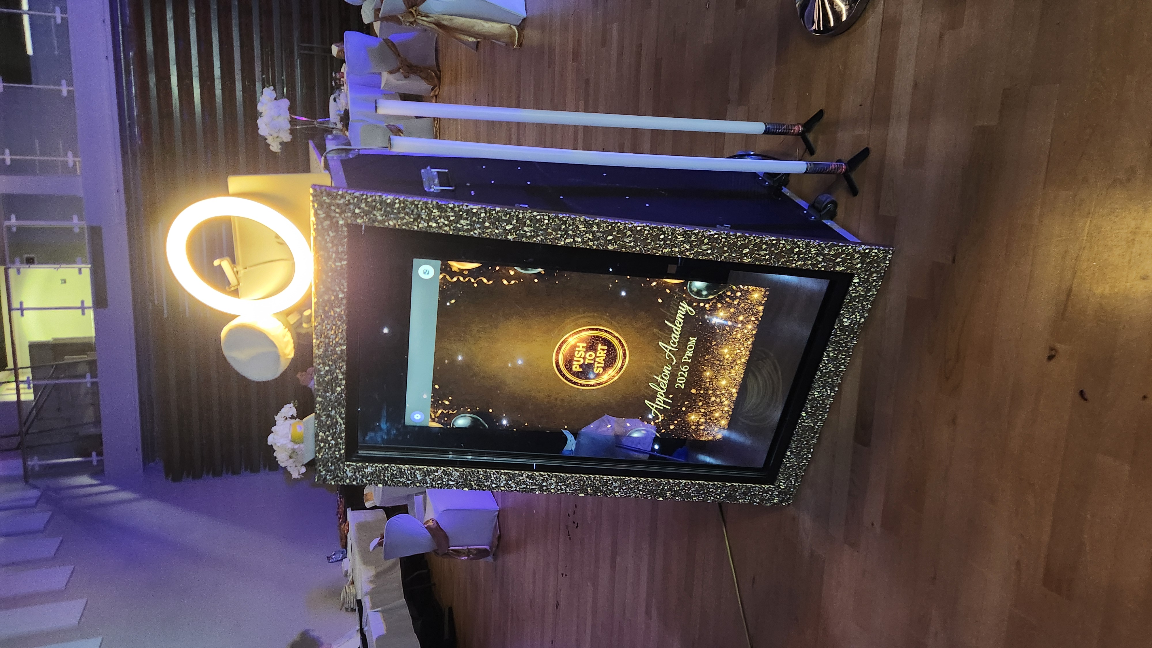 Glitter-framed photo booth with glowing ring light, on hardwood floor amid white-draped prom tables and chairs in cool purple ambient light.