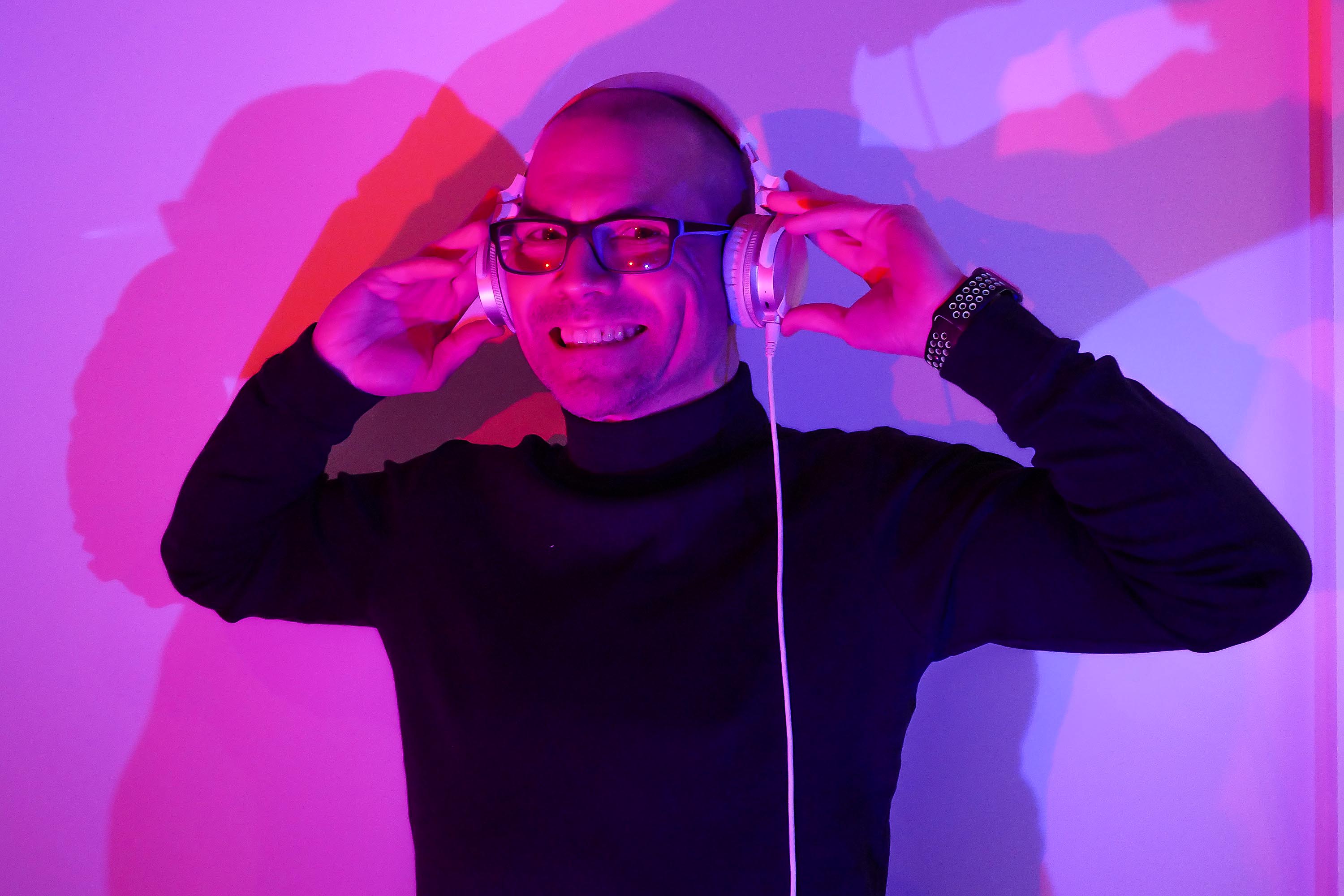 A smiling DJ in glasses and headphones poses under vivid magenta-blue lighting, black turtleneck, clean modern event atmosphere.