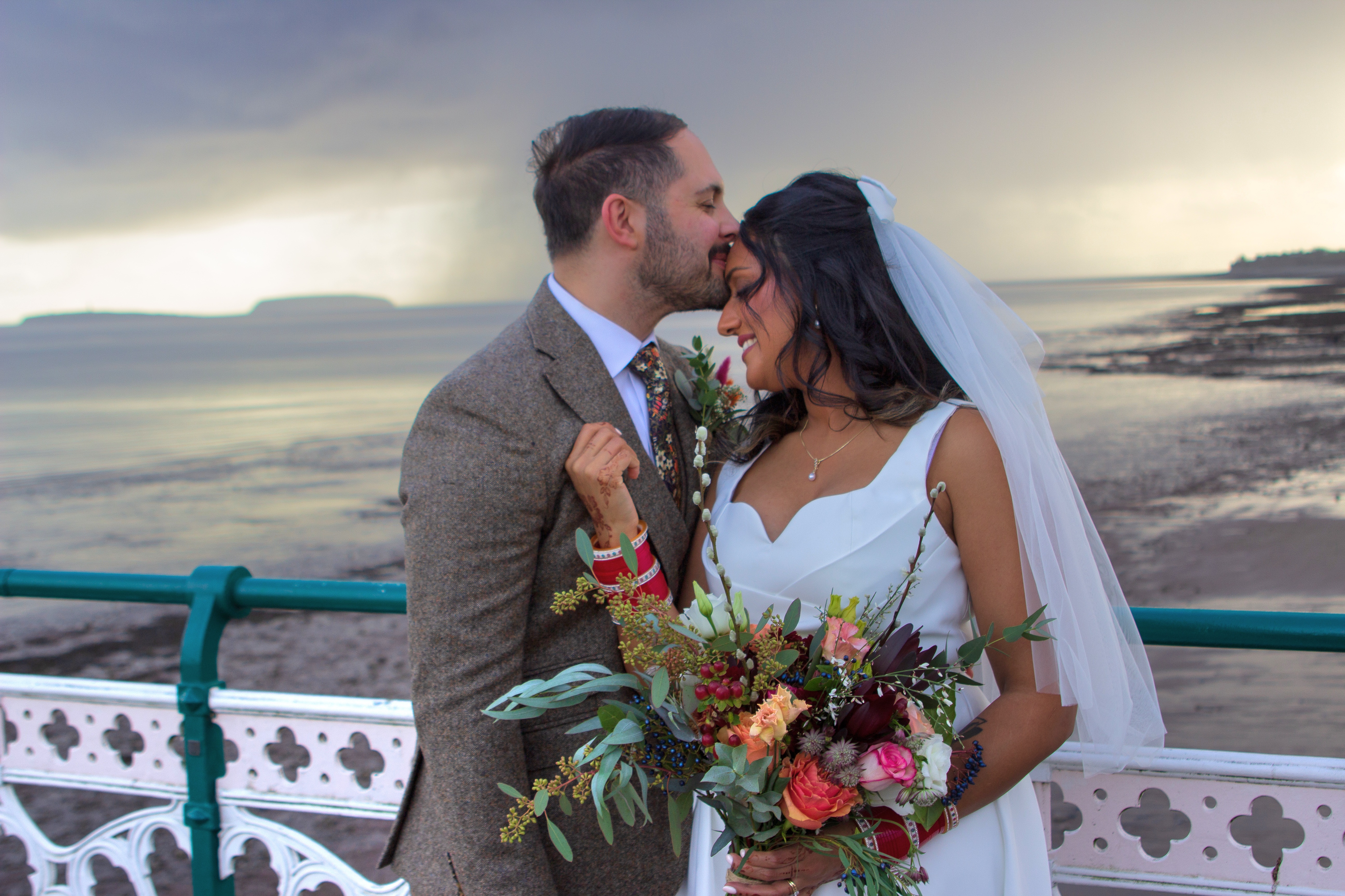 A couple embraces on a windswept beach, surrounded by dramatic skies. The bride holds a vibrant bouquet with rich red and pink blooms, radiating romance.