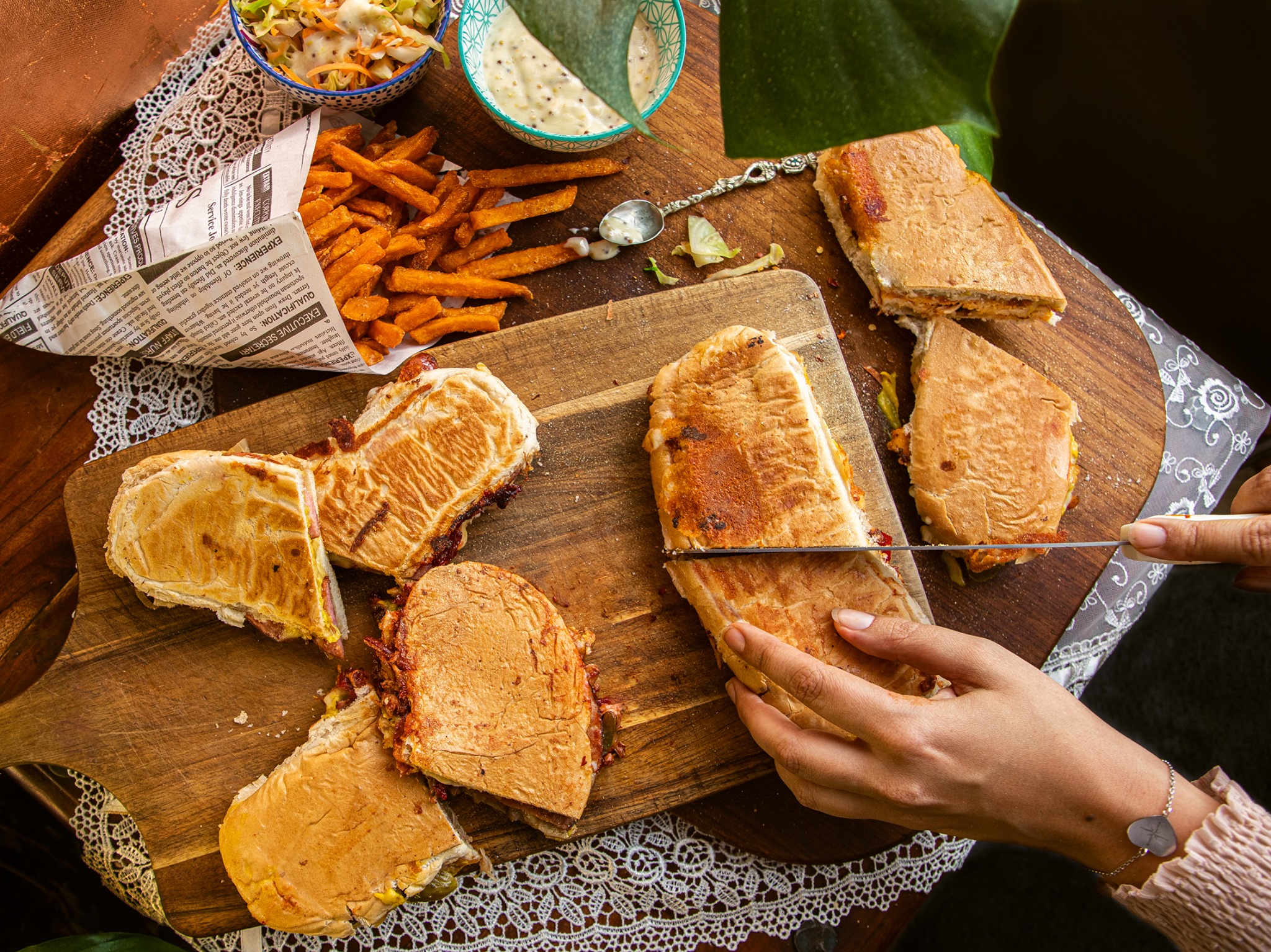 A wooden cutting board displays grilled sandwiches and sweet potato fries in a newspaper cone, surrounded by colorful cole slaw and dipping sauce, all set under warm, inviting light.