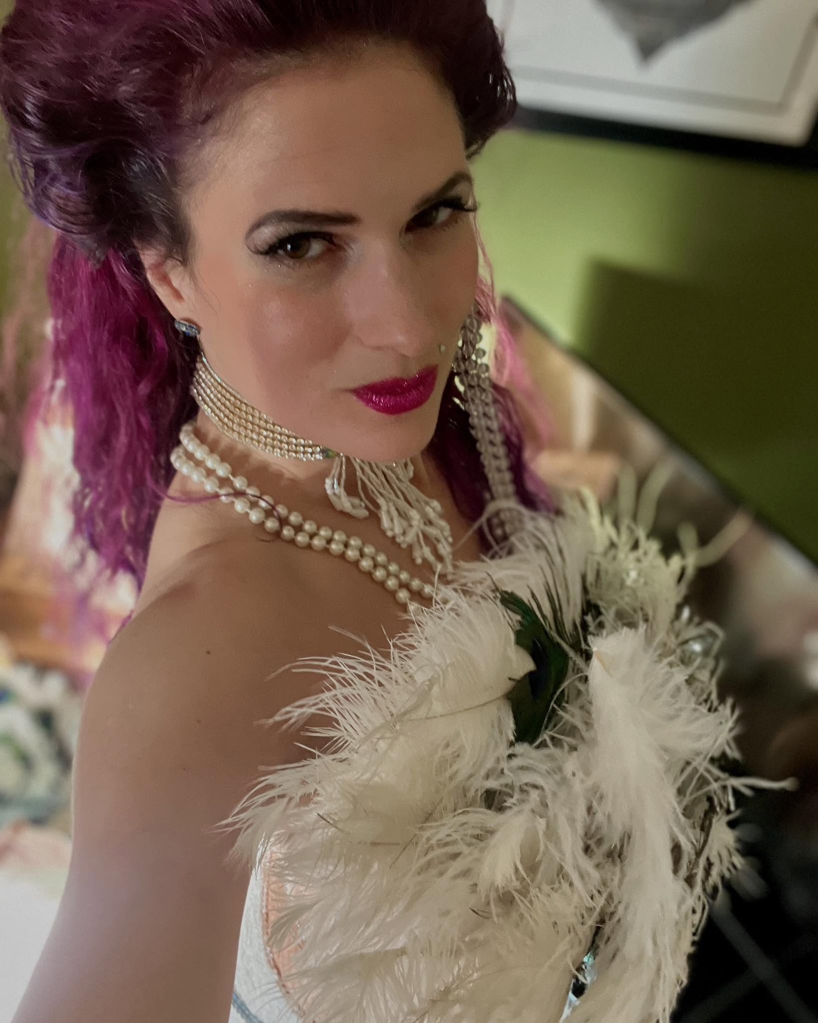 Glamorous close-up portrait of a woman with magenta hair, pearl necklace, and feathered white couture, softly lit indoors.