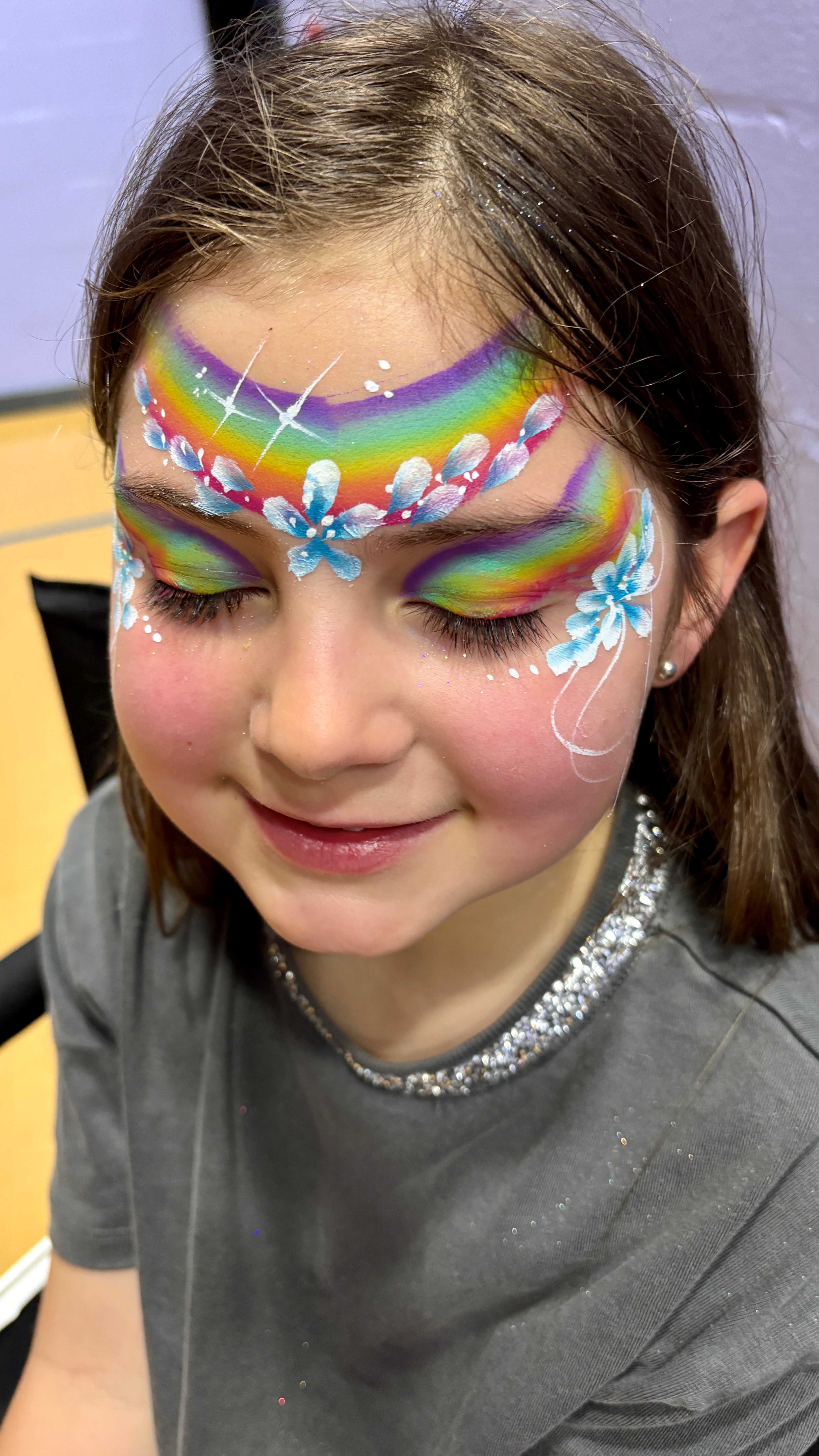 Close-up of a smiling child with rainbow face painting and white-blue floral accents, under bright indoor light, showcasing vibrant artistry.