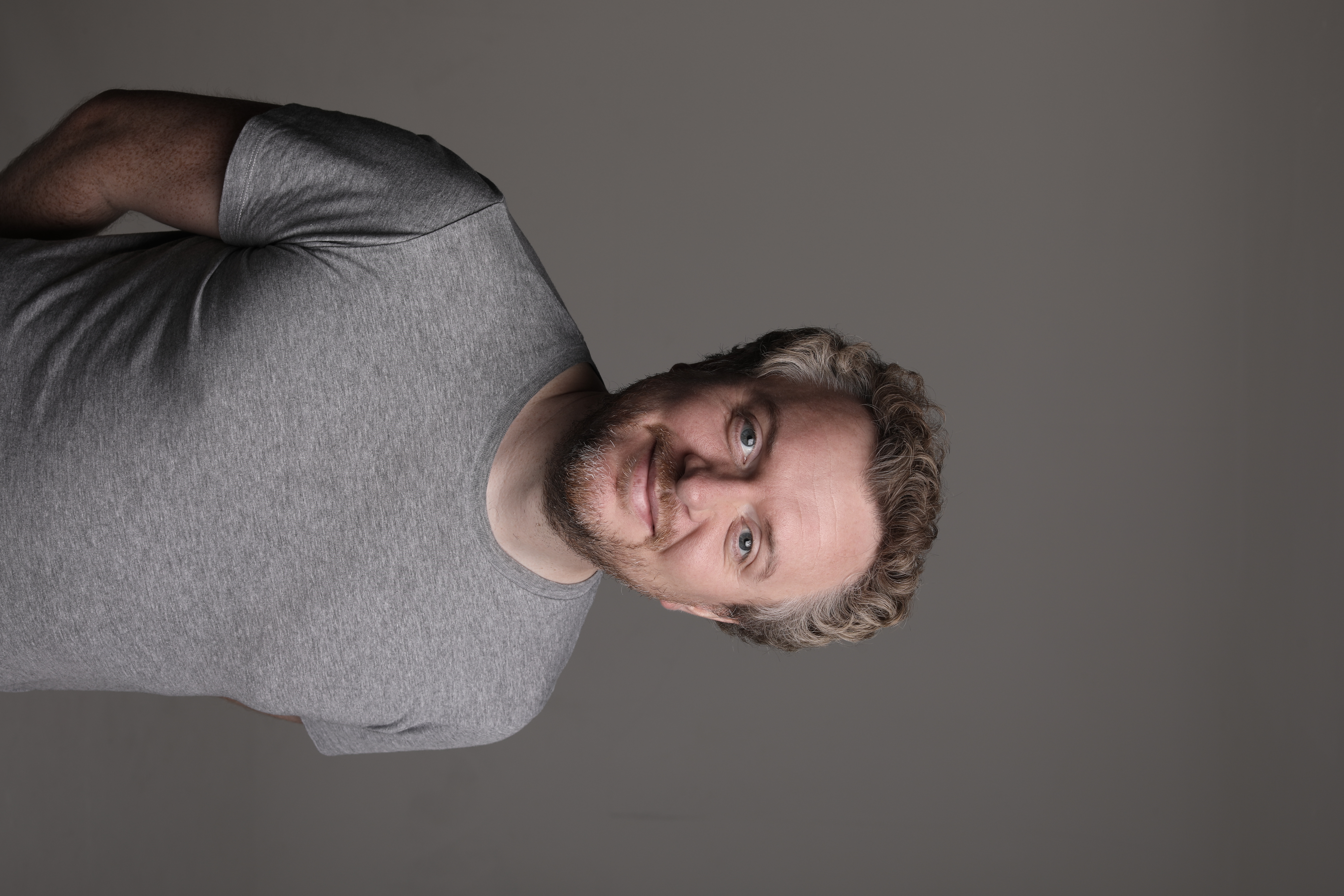 A cheerful man with curly hair and a beard wears a simple gray t-shirt against a soft, neutral backdrop, exuding a friendly, approachable vibe.