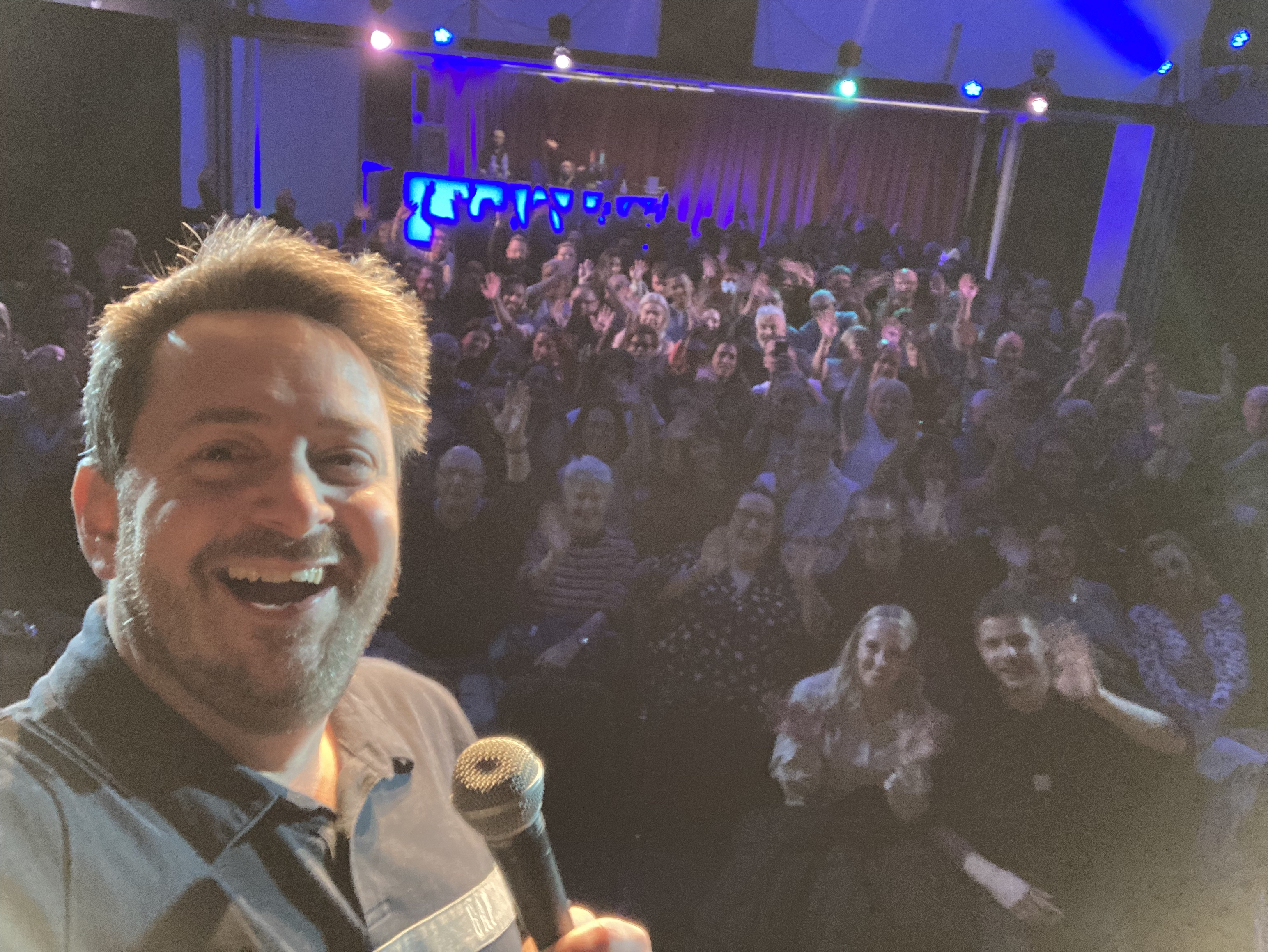 Smiling host with microphone selfies a packed, cheering audience under blue-purple stage lights, lively event-catering atmosphere.