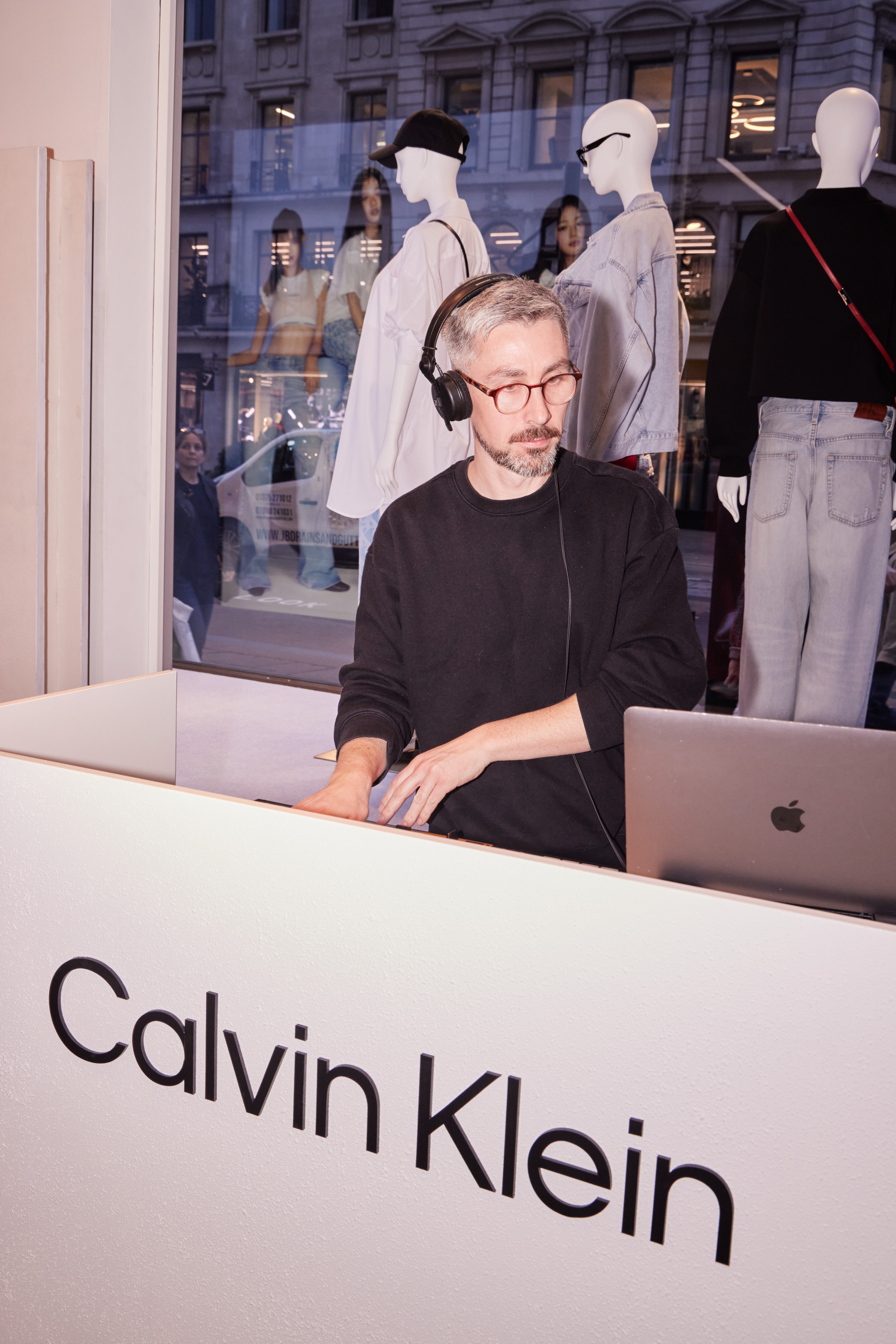A stylish shop scene shows a man in black headphones working at a sleek white counter, while trendy mannequins showcase modern apparel.