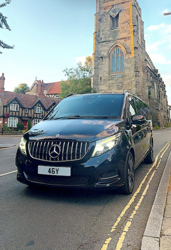 A sleek black Mercedes van is parked on a quiet street, with a historic stone church towering in the backdrop, illuminated by soft daylight.