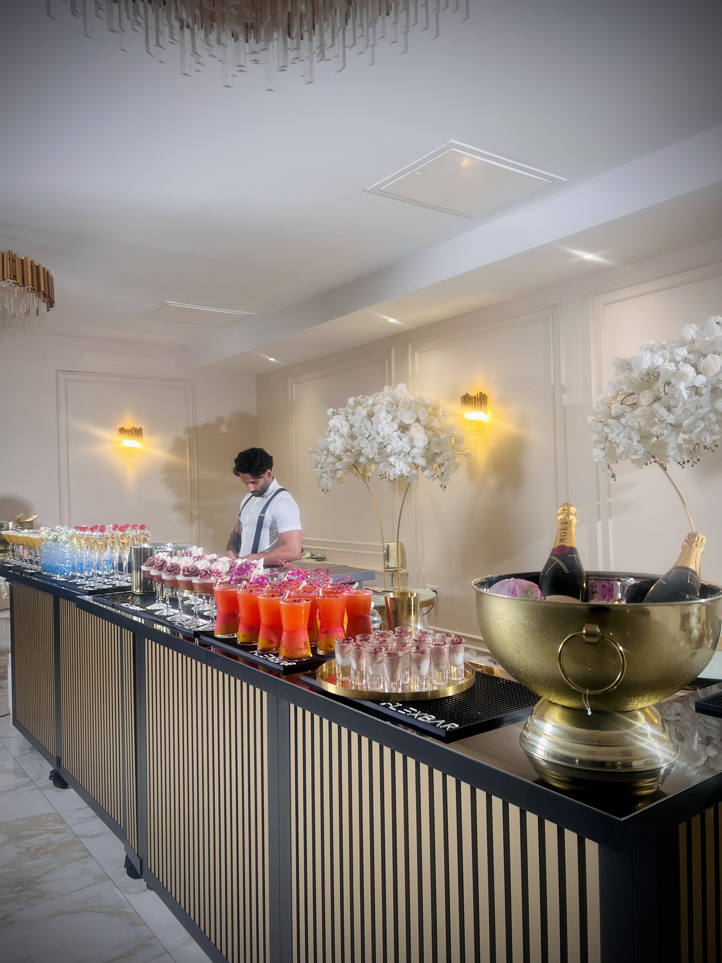 A sleek bar features vibrant cocktails in elegant glassware, with white floral arrangements and warm lighting creating an upscale atmosphere.