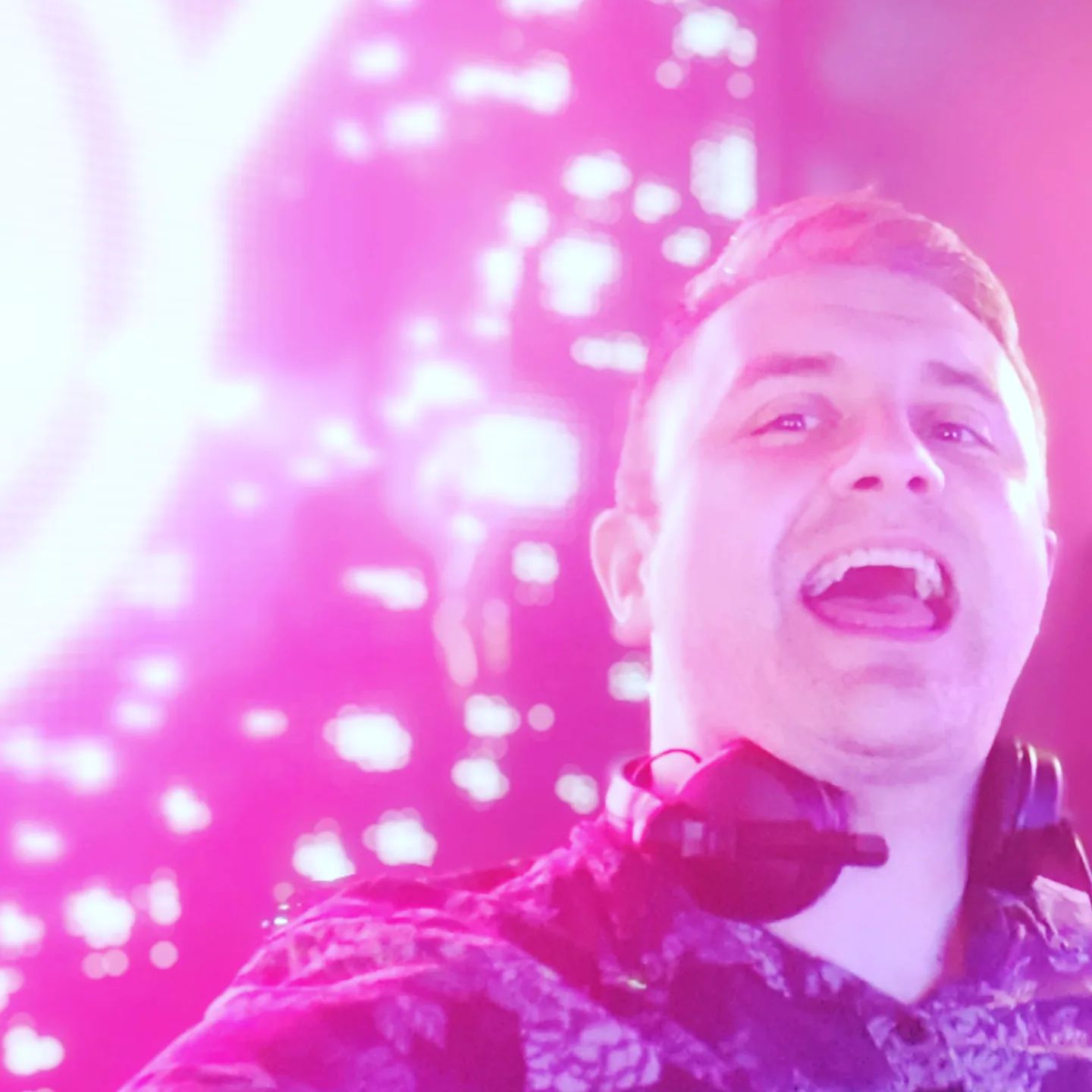 A lively DJ smiles widely, illuminated by vibrant pink and purple lights, creating an electrifying atmosphere perfect for an energetic event.
