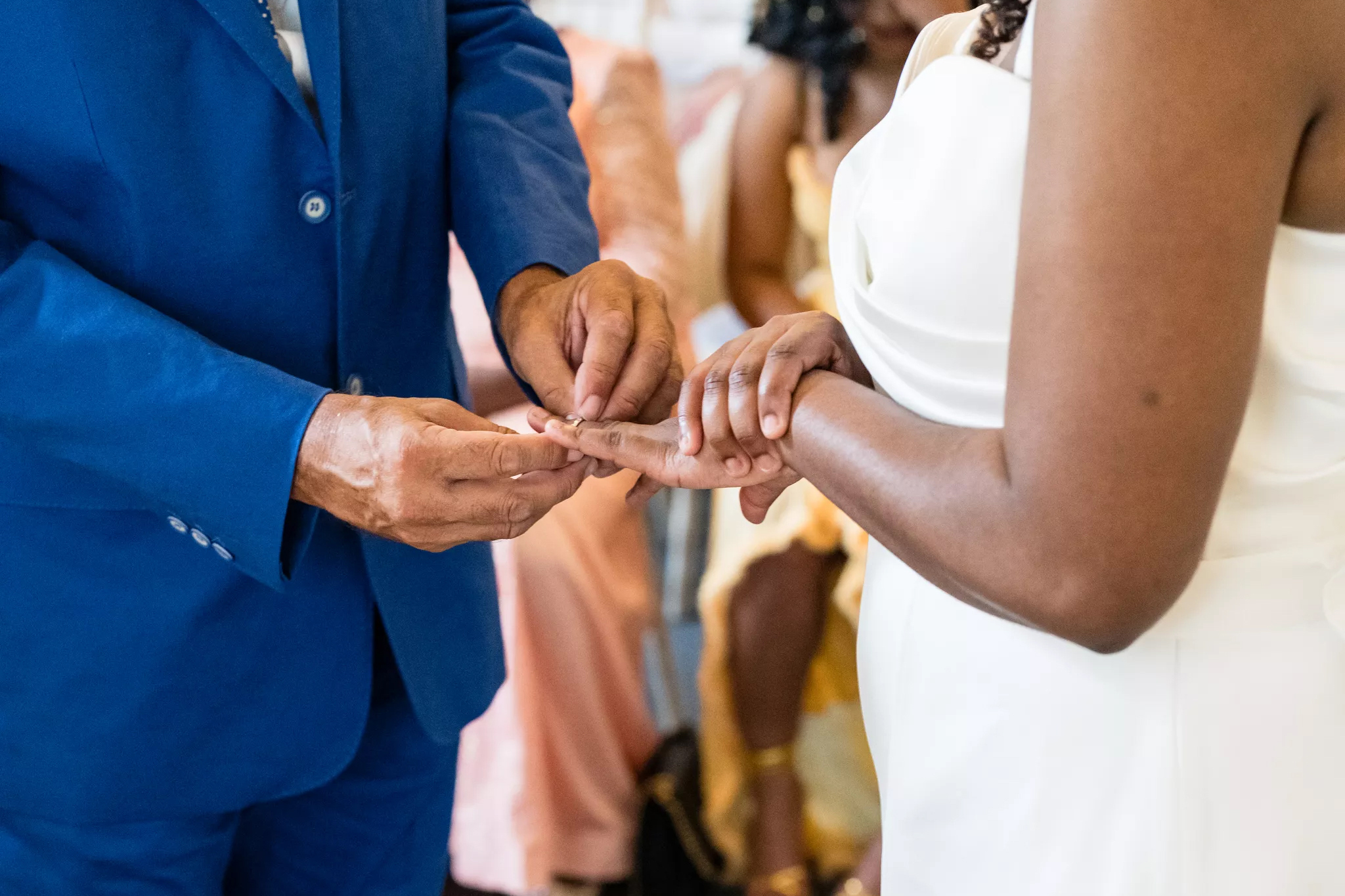 At a warmly lit wedding, a groom in a royal-blue suit places a ring on a bride’s hand, with blurred guests and elegant ceremony focus.