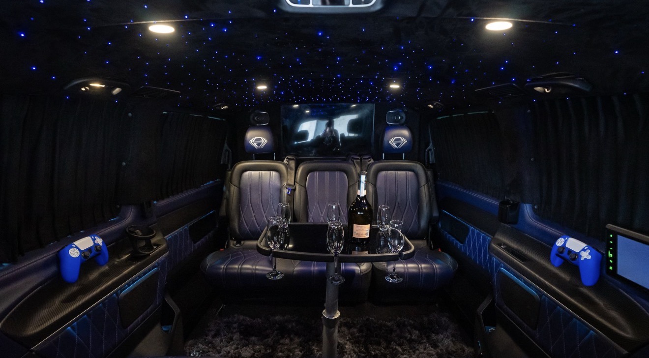 Inside a luxurious van, plush navy seats face a black table holding a champagne bottle and crystal flutes, under starry blue lighting.