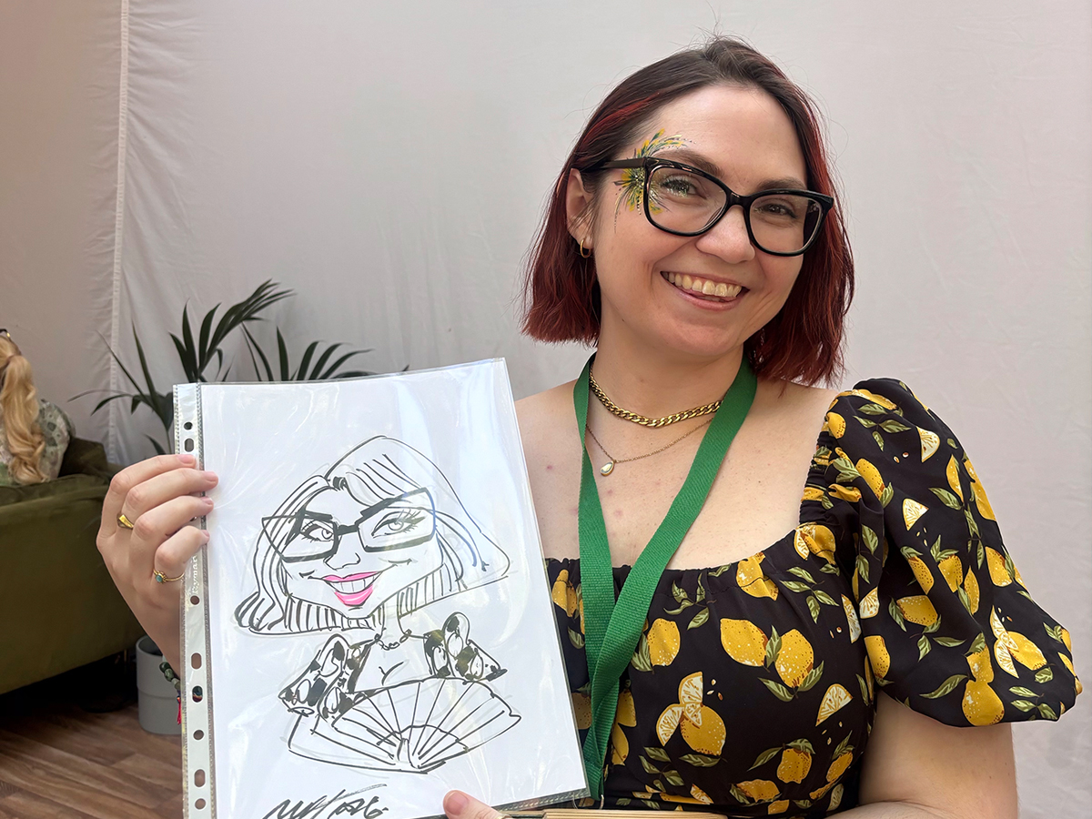 Smiling caricature artist at a bright catering event, holding a crisp sketch in a plastic sleeve; lemon-print dress, polished glasses, warm atmosphere.
