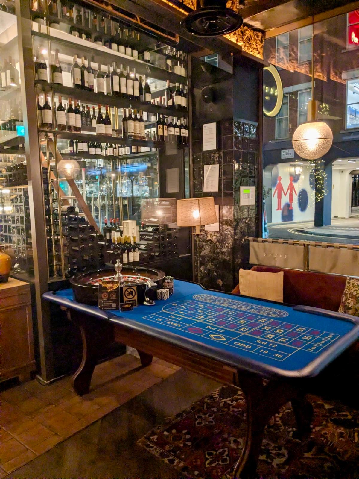 A moody bar features a roulette table with a vibrant blue felt, surrounded by polished wood, cozy cushions, and a stunning wine display.