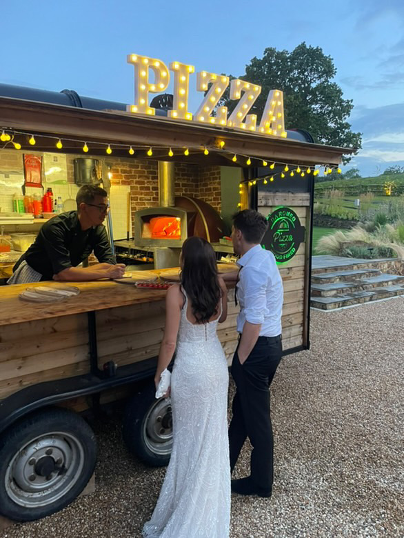 Twinkling pizza catering trailer with wood counter, glowing oven, and polished service at dusk as guests order outdoors.