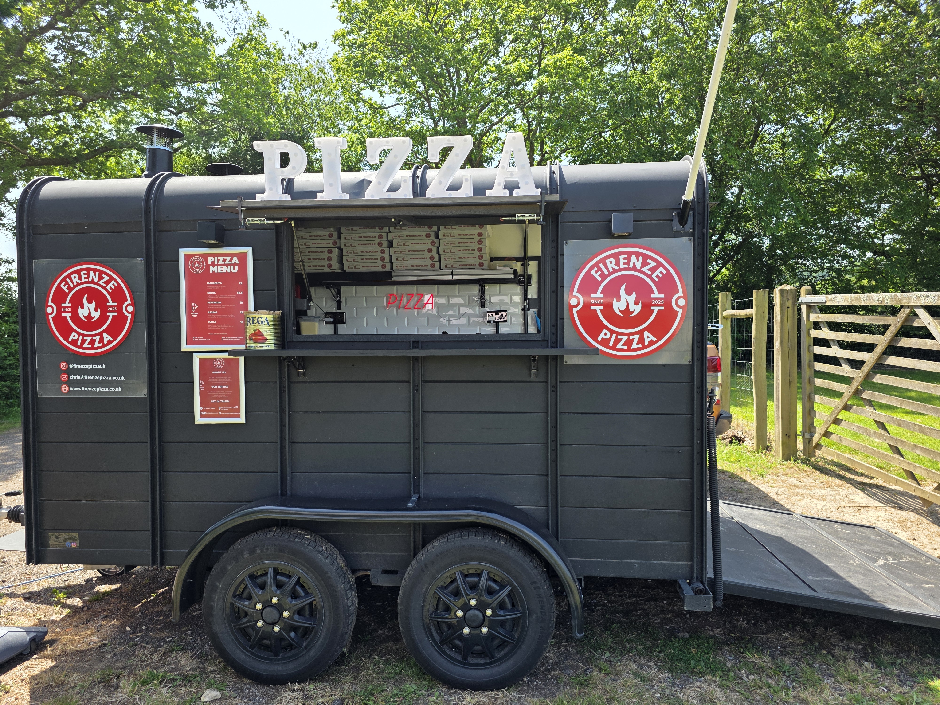 A vintage black pizza trailer displays a vibrant red sign, illuminated "PIZZA" letters, and a menu of gourmet options. The inviting setup enhances outdoor catering vibes.