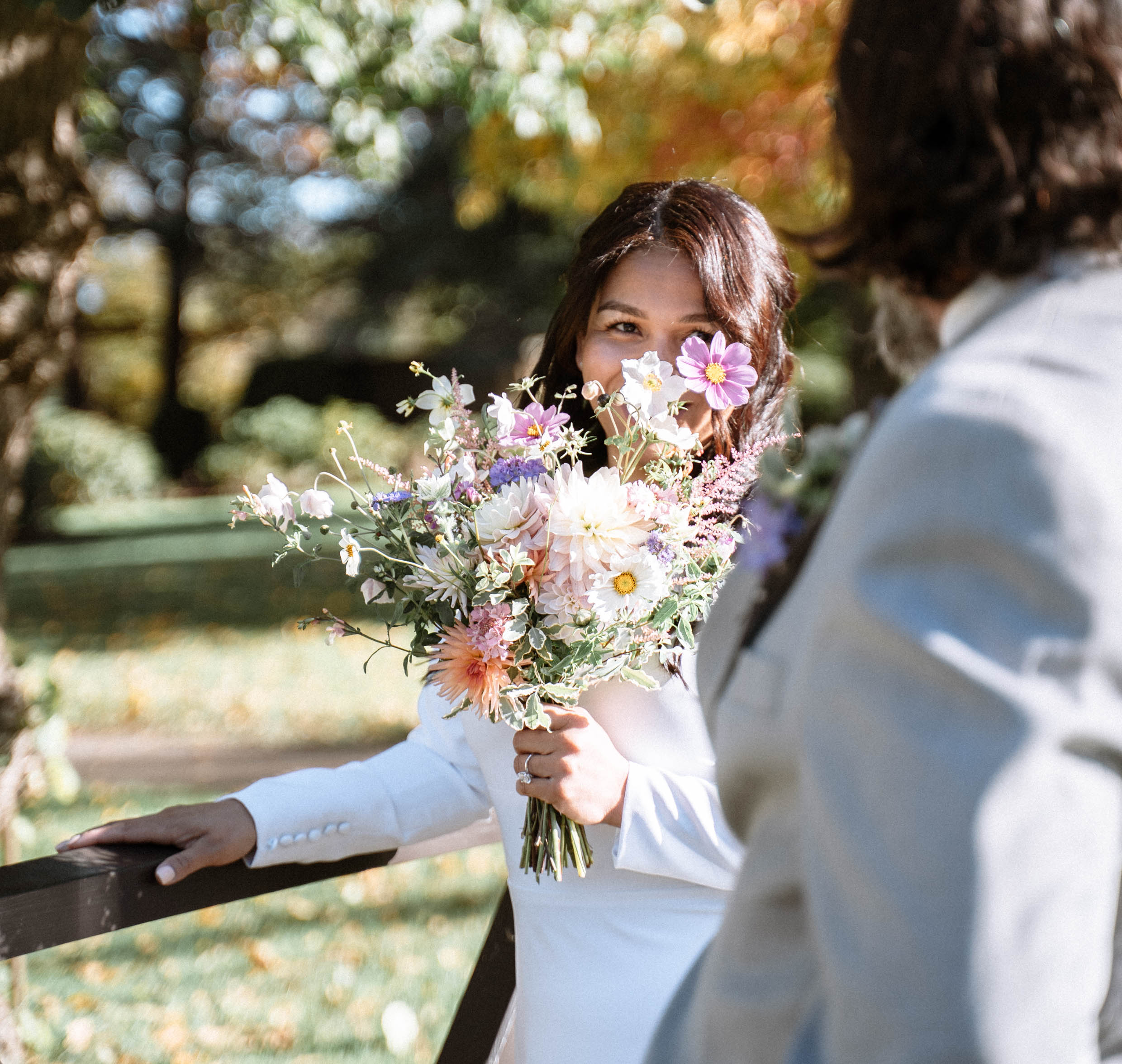 A bride in a flowing white gown holds a vibrant bouquet filled with pink and purple flowers, smiling in a sunlit garden, exuding joy.