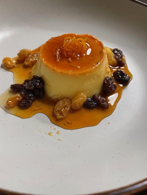 A glossy flan topped with orange zest sits elegantly on a white plate, surrounded by glistening golden raisins in a rich caramel sauce.