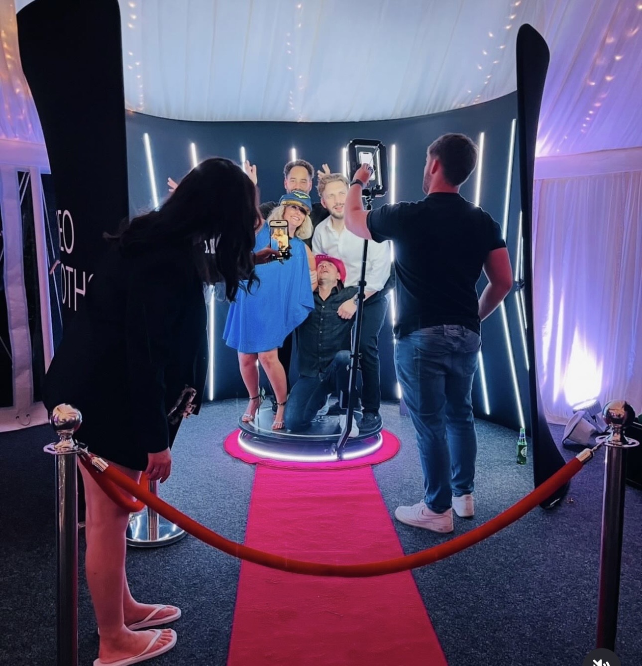 Guests pose on a glowing 360 photo booth platform, framed by LED strips, a red carpet, and velvet ropes under soft white drapes.