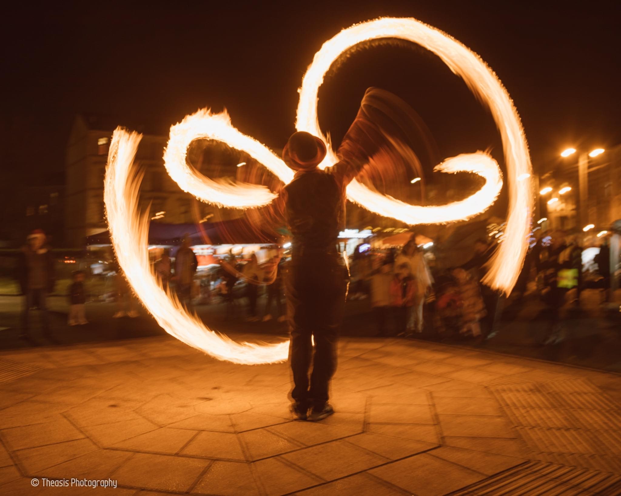 A performer spins fire poi in swirling arcs, illuminated by warm streetlights, captivating an audience in a lively nighttime market scene.