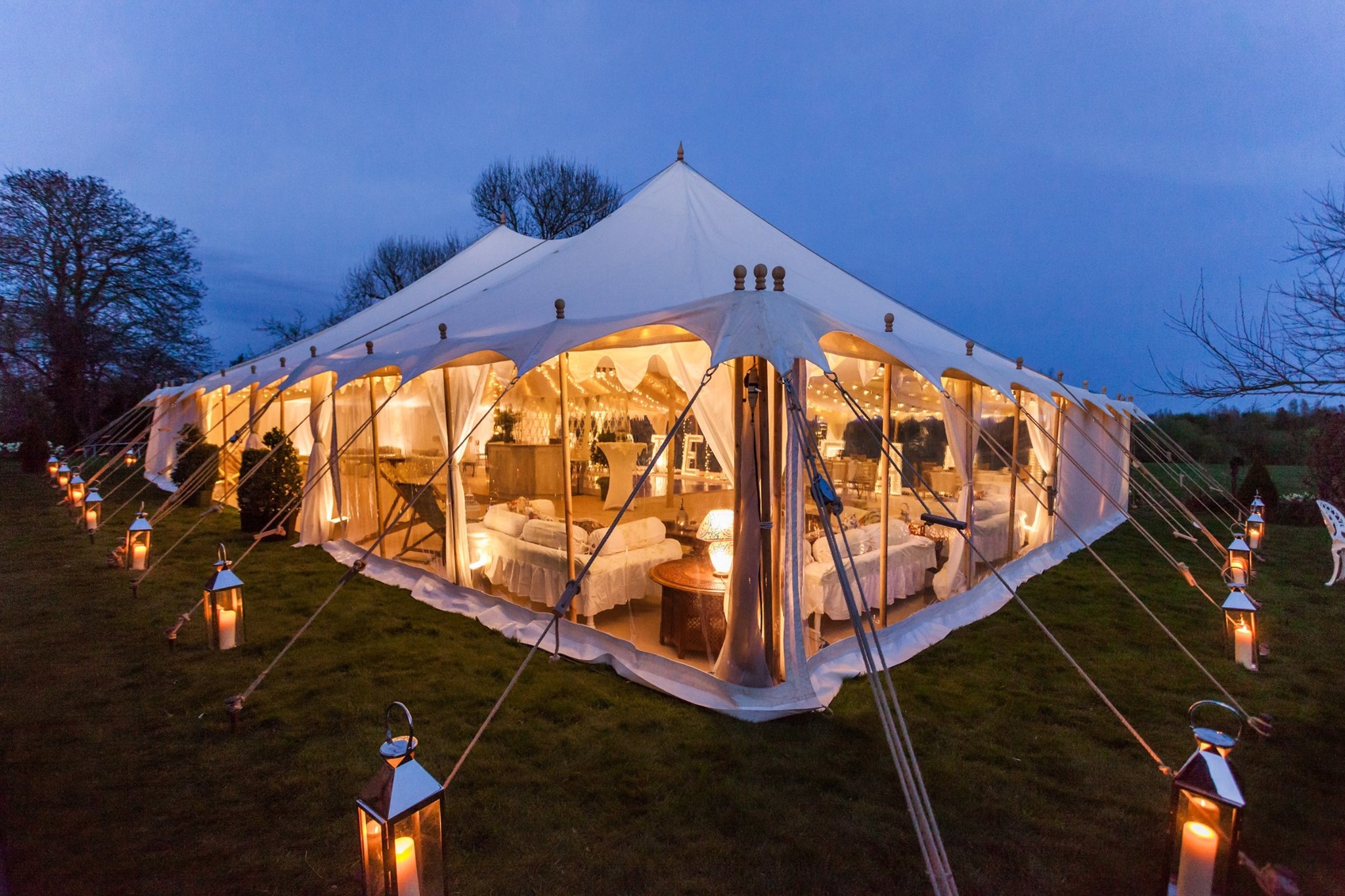 Elegant white marquee tent with warm lanterns, draped linen, and polished event seating, glowing at dusk for upscale catering.