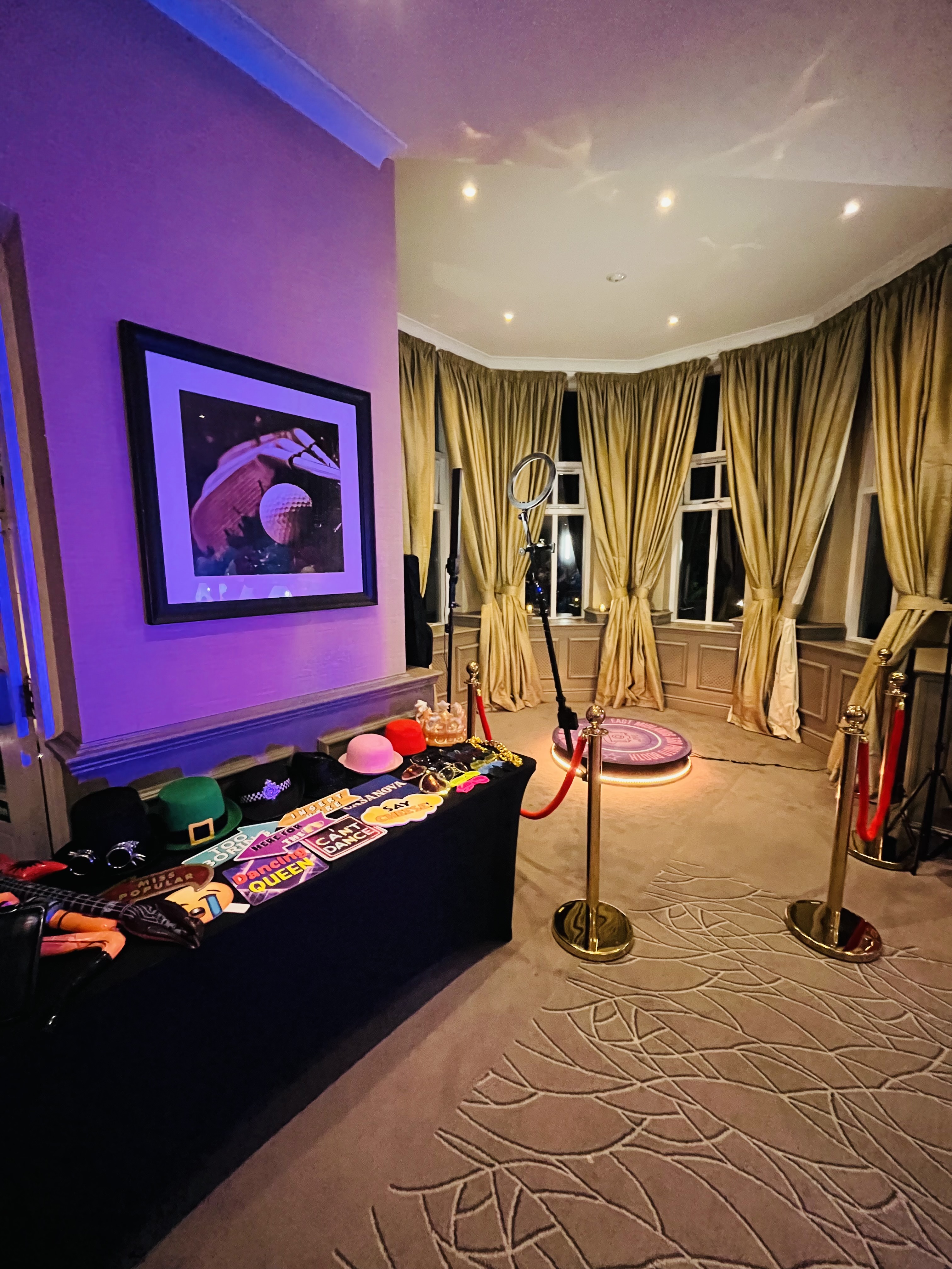 Purple-lit photo booth with ring light, gold drapes, velvet rope, and hat props on a black catering table in a chic lounge.
