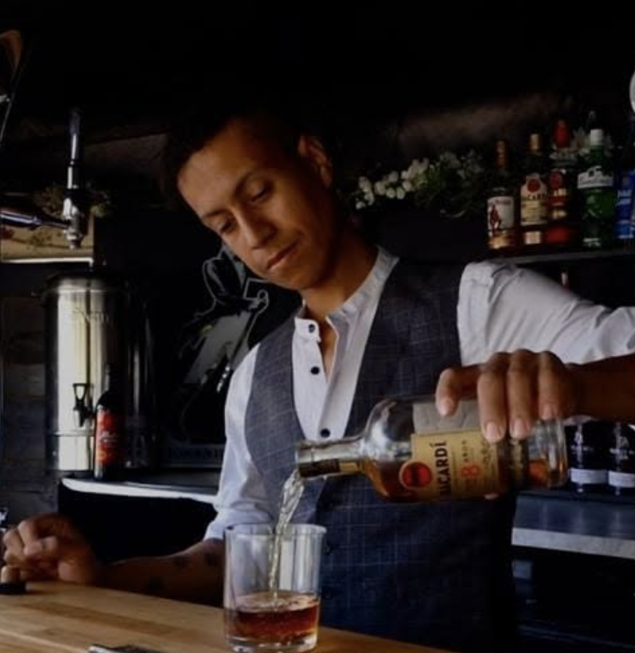 Bartender in a crisp vest pours Bacardi into a glass at a rustic wood bar, with polished bottles and warm moody lighting.