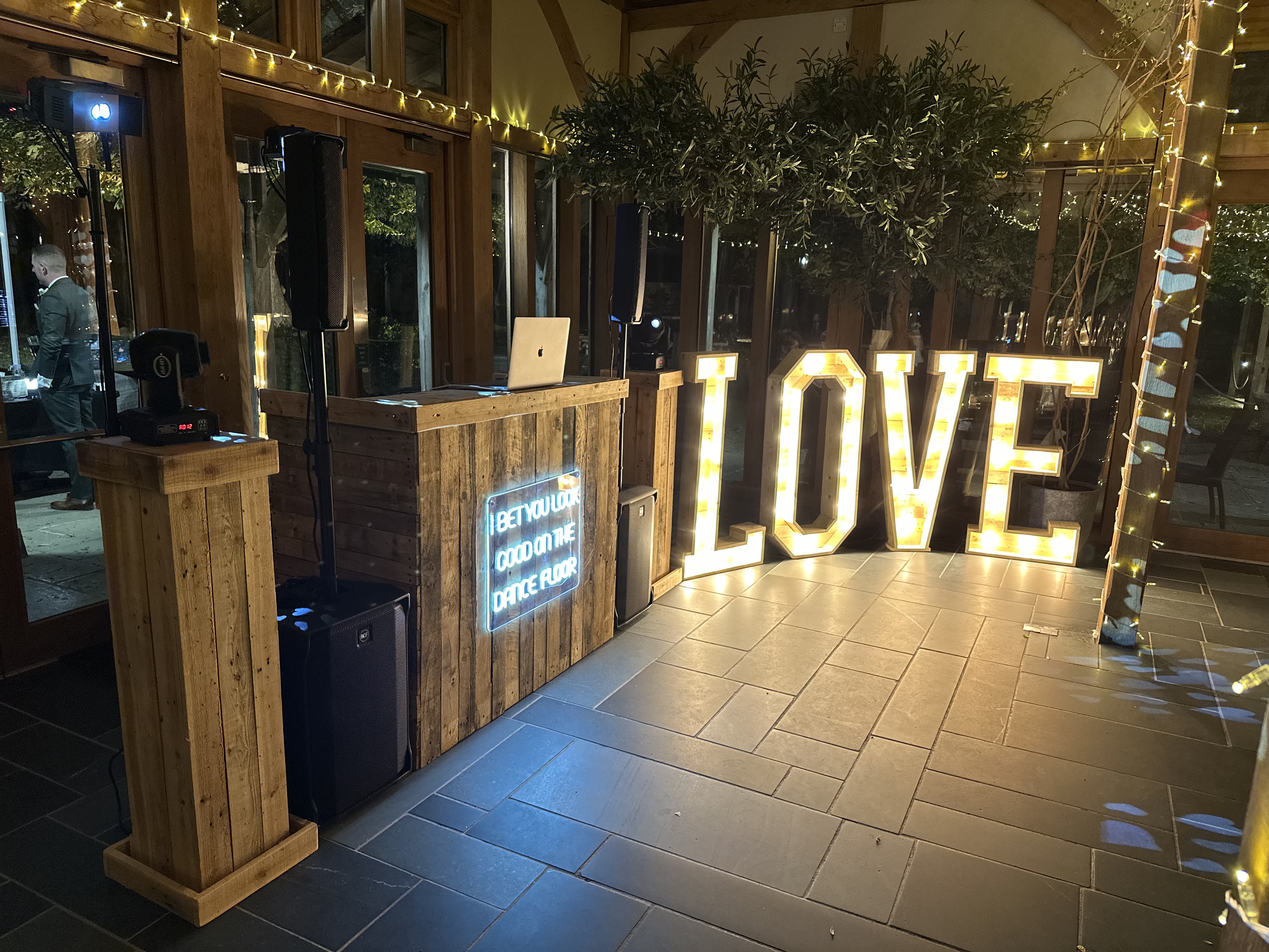 A rustic wooden DJ booth with decorative lights showcases a vibrant "LOVE" sign, illuminated warmly against a sleek stone floor.