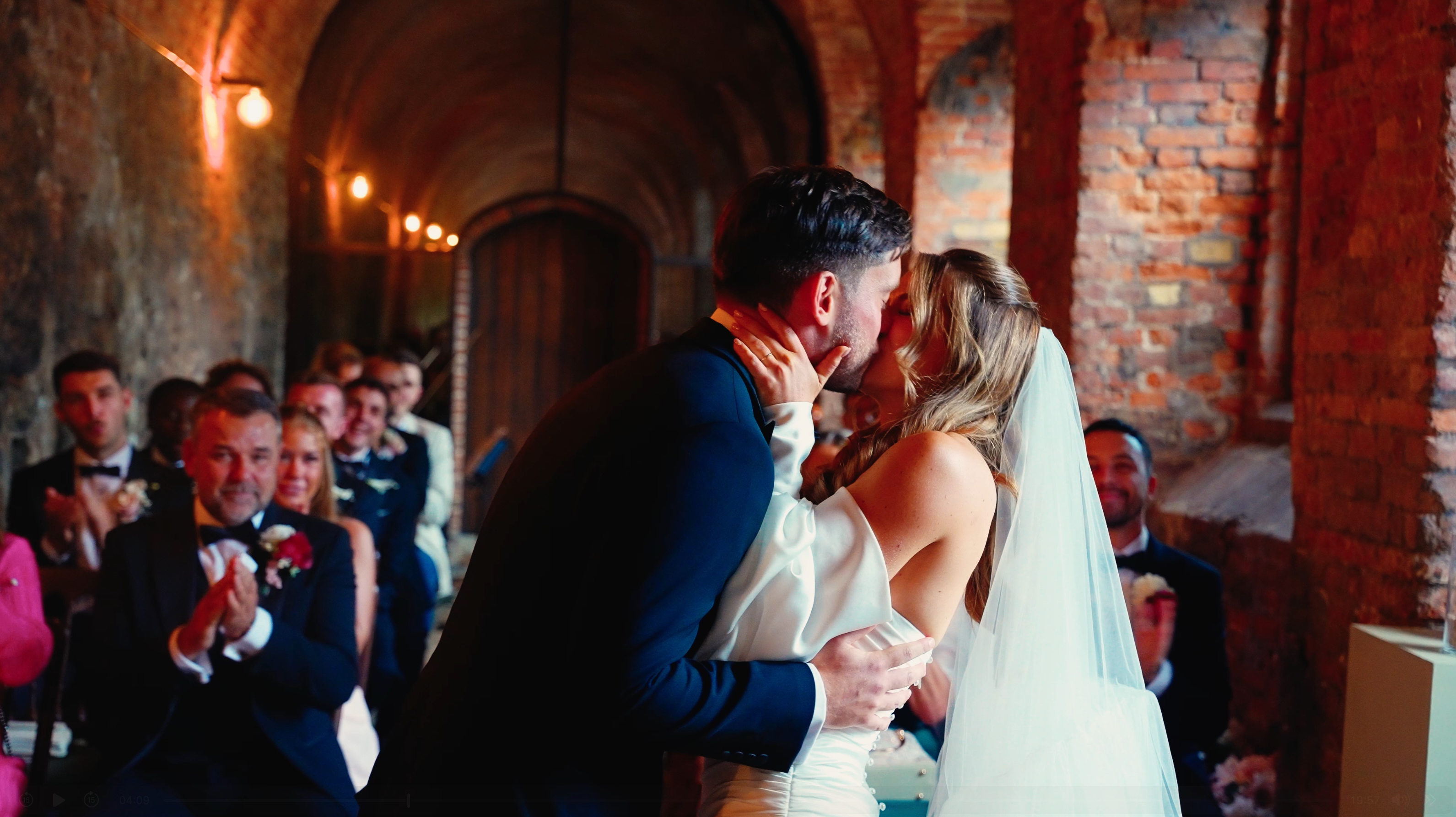 Bride and groom kiss in a brick-vaulted venue with warm candlelight, while suited guests applaud in a romantic wedding ceremony.